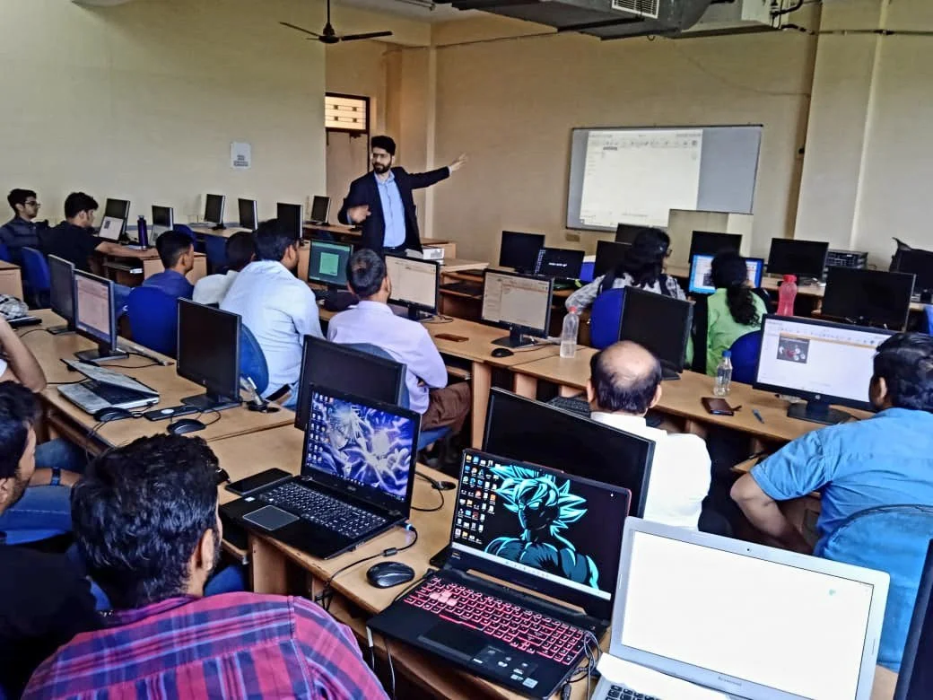 A classroom setting with students using computers, and a teacher pointing at a screen.
