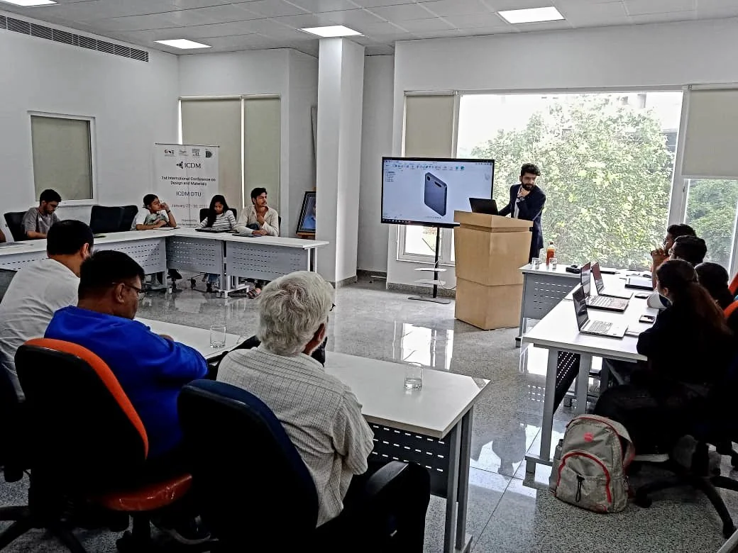 A business meeting with people sitting around conference tables, a presenter at a podium, and a large screen displaying a presentation.