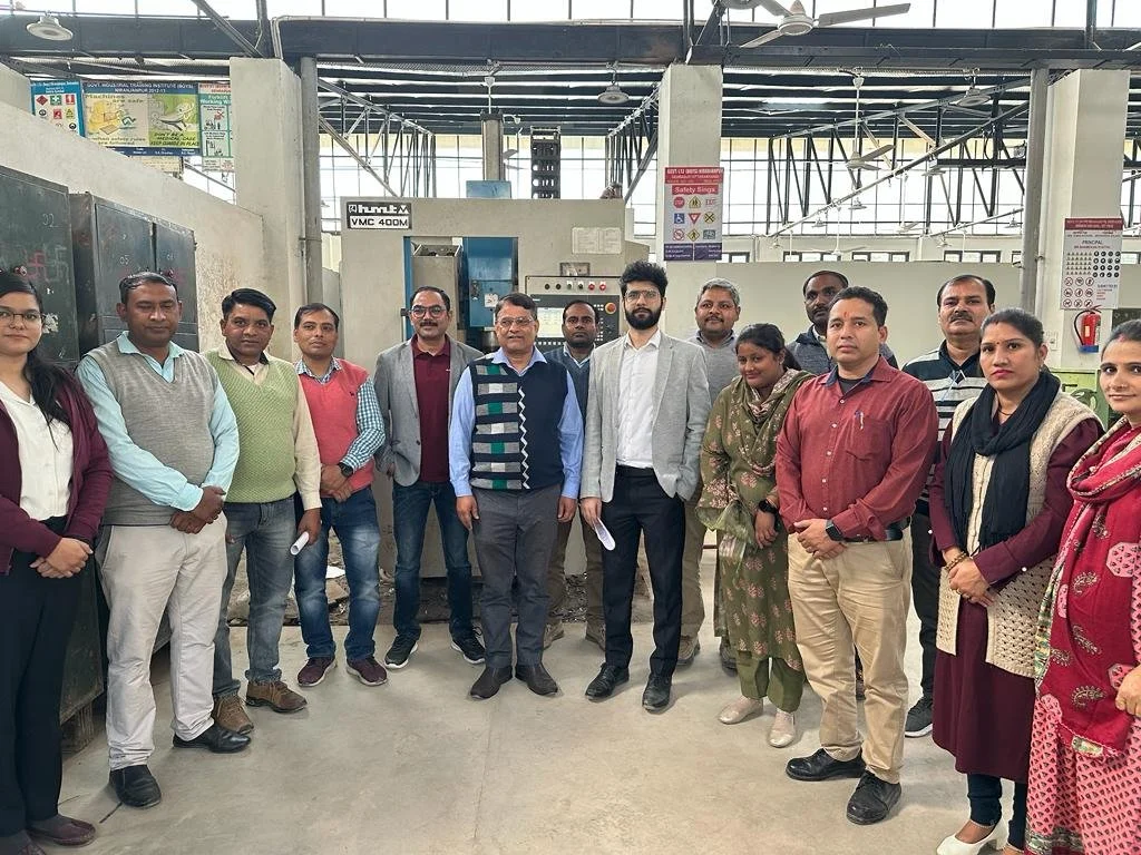 A group of people standing in a row inside an industrial or workshop setting, with machinery and safety signs visible in the background. They appear to be posing for a group photo.