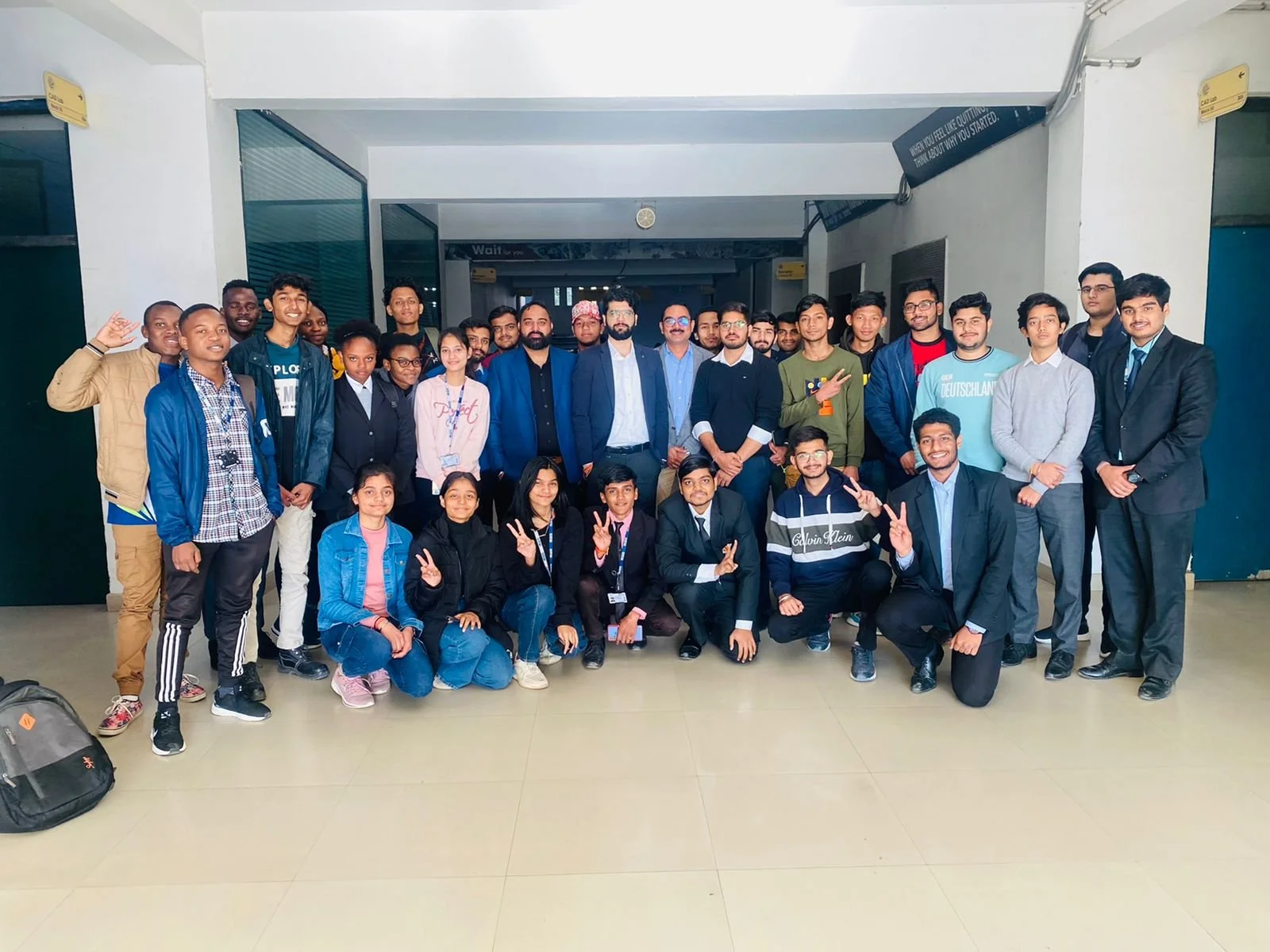A group of people standing and kneeling in a hallway, posing for a photo. Some are wearing casual clothes while others are in formal attire. The group includes men and women of various ages, with some making peace signs with their hands. There is a b