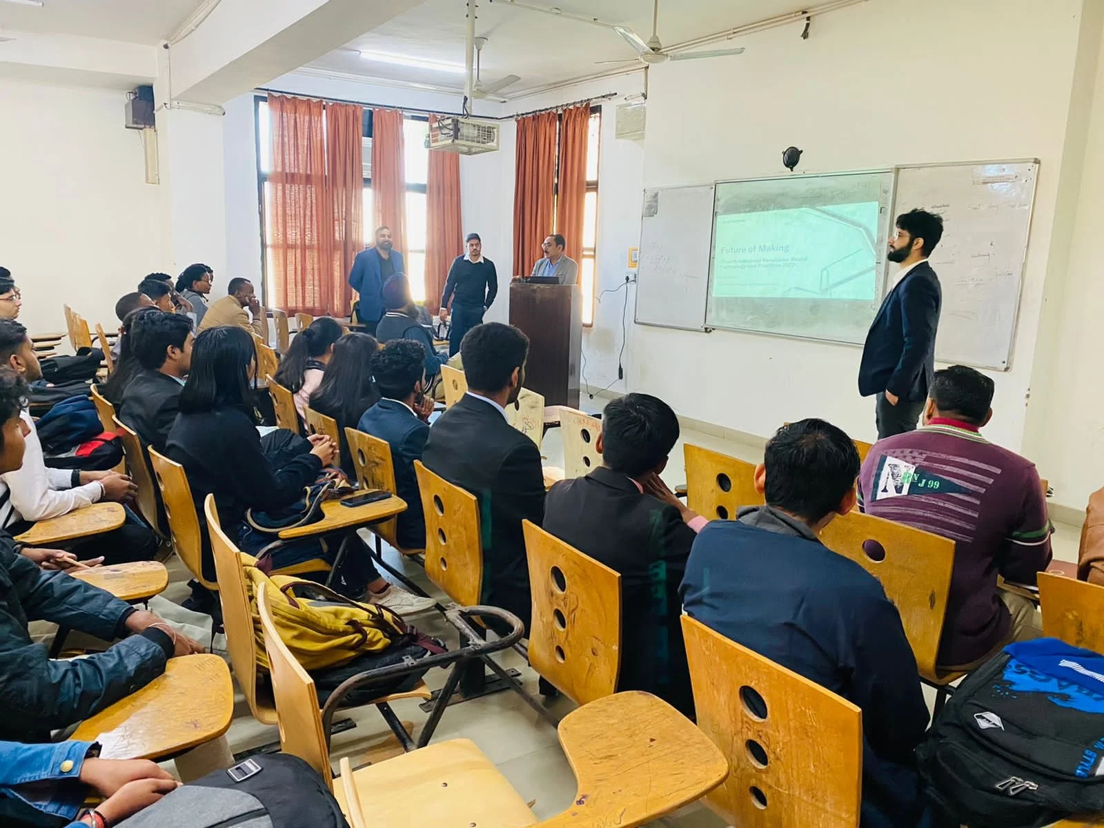 Classroom with students and a presenter giving a lecture, projector screen displaying a presentation with several people standing at the front.