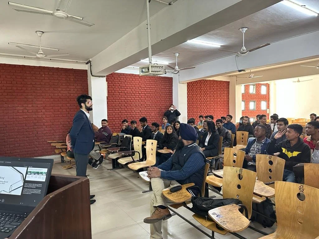 A classroom setting with a male speaker addressing a group of students seated in chairs. The room has red brick walls and a laptop on a podium displays a presentation.