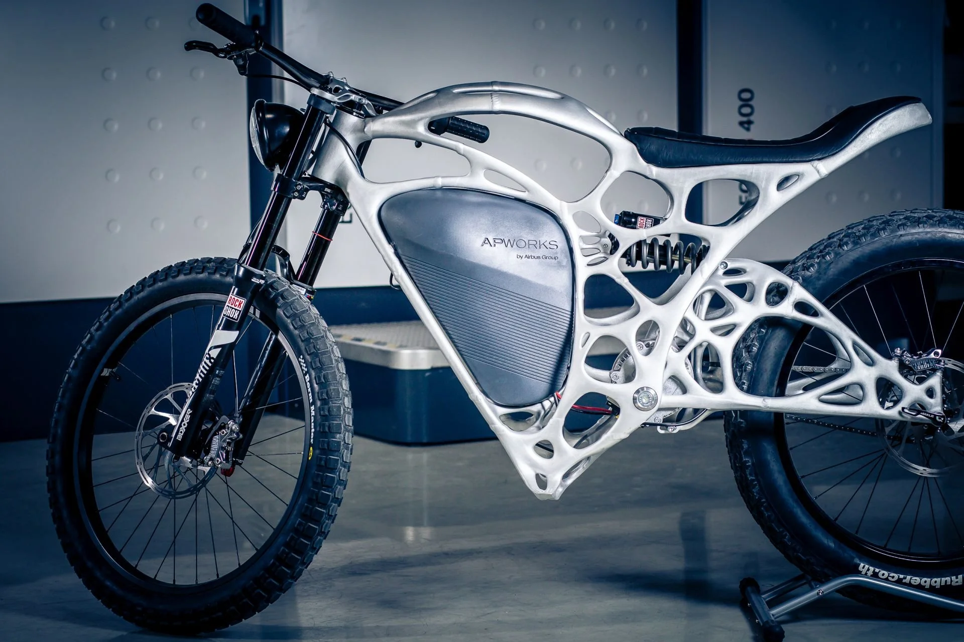 Futuristic electric bicycle with advanced lightweight frame design, labeled APWorks by Airbus Group, standing indoors.