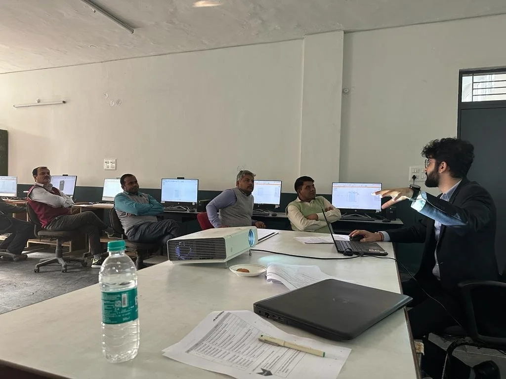 A group of people in a computer lab attending a presentation. One person is gesturing while speaking, and others are sitting at desks with computers. A bottle of water, papers, and a laptop are on the table.