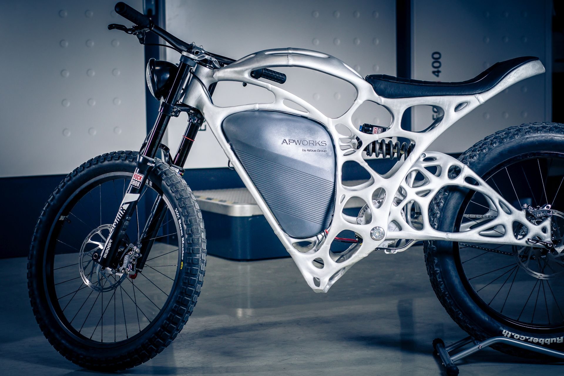 Futuristic electric bicycle with advanced lightweight frame design, labeled APWorks by Airbus Group, standing indoors.