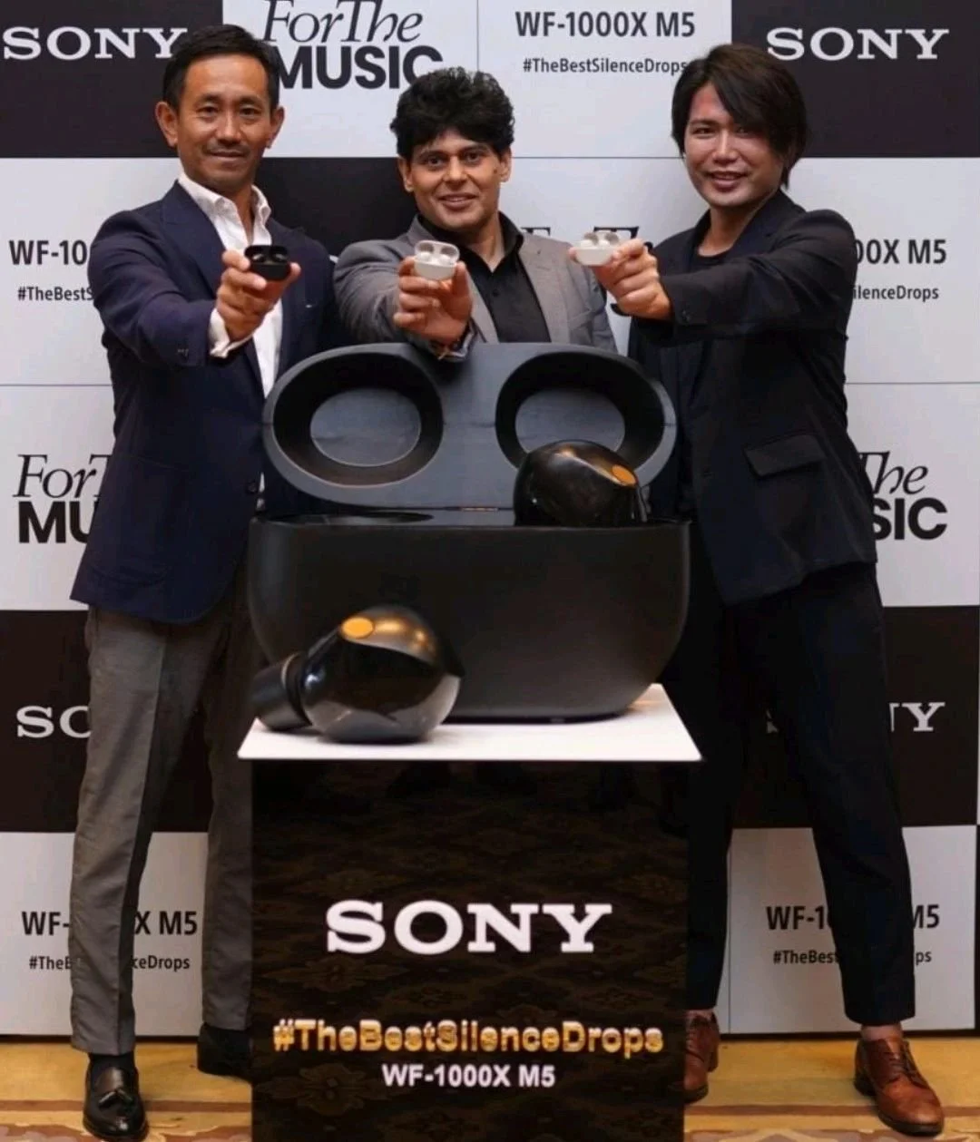 Three men in suits holding Sony earbuds in front of a backdrop with "Sony," "For the Music," and "WF-1000X M5 #TheBestSilenceDrops" text, showcasing audio products.