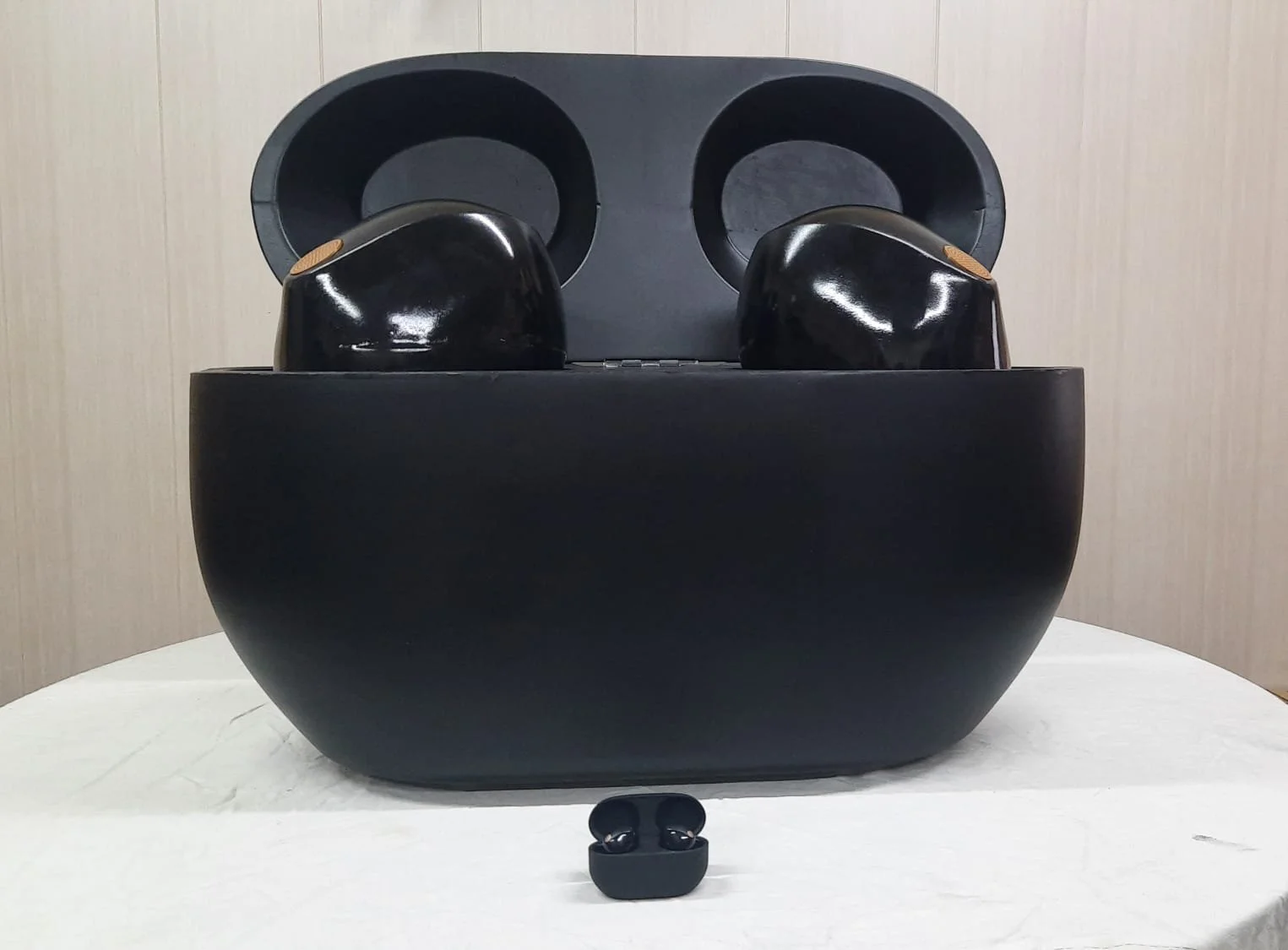 Black wireless earbuds in a charging case, with a smaller pair in front for size comparison, on a light surface against a wooden background.