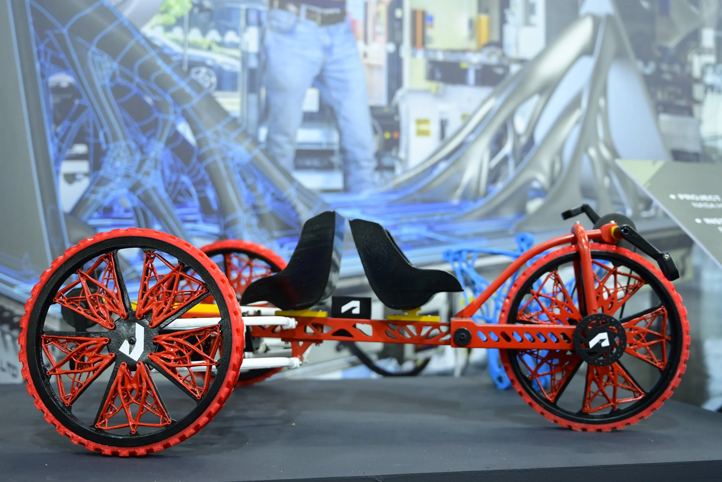 Futuristic tricycle model with red and black wheels, dual seats, and a sleek design on display. Computer-generated backgrounds visible.