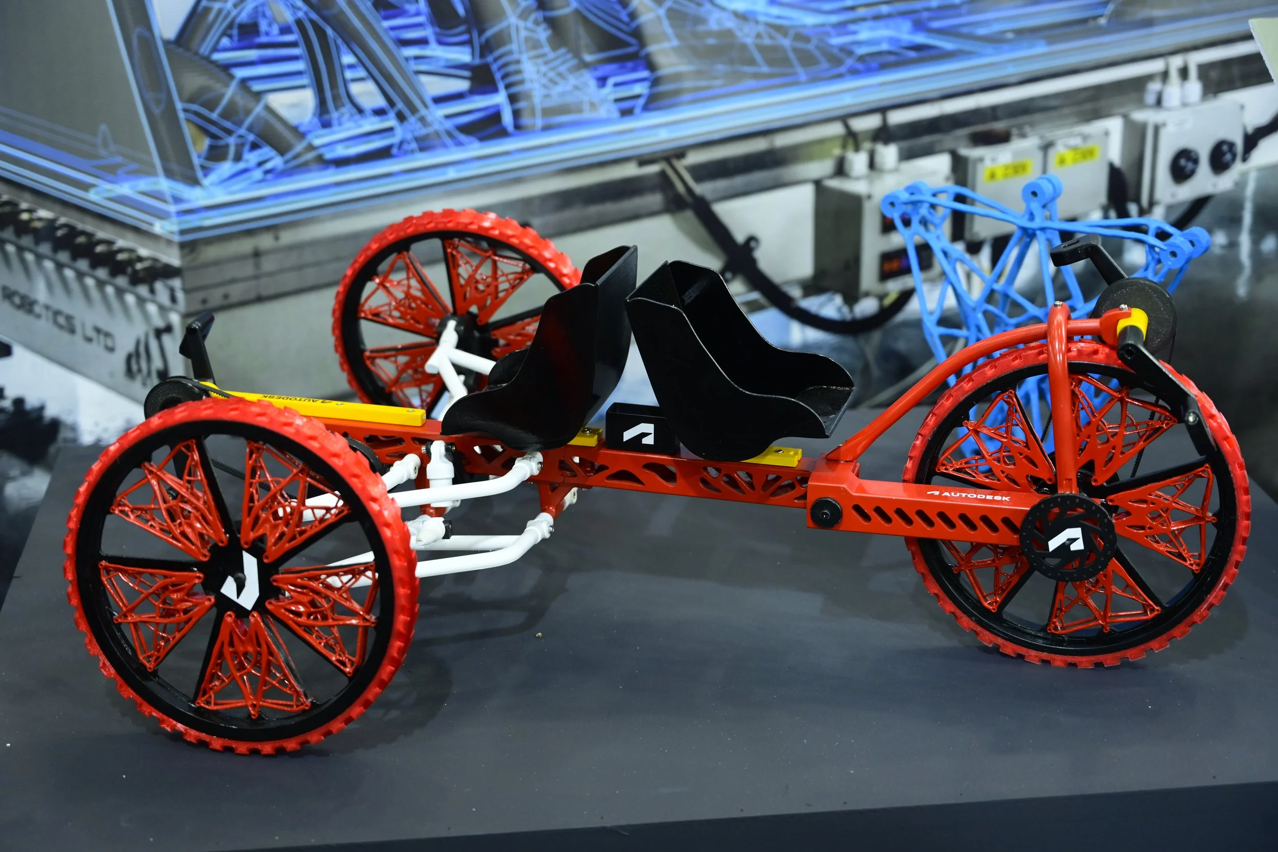 A model of a futuristic four-wheeled vehicle with red wheels and a lightweight frame displayed on a table. The background features industrial machinery and blueprints.
