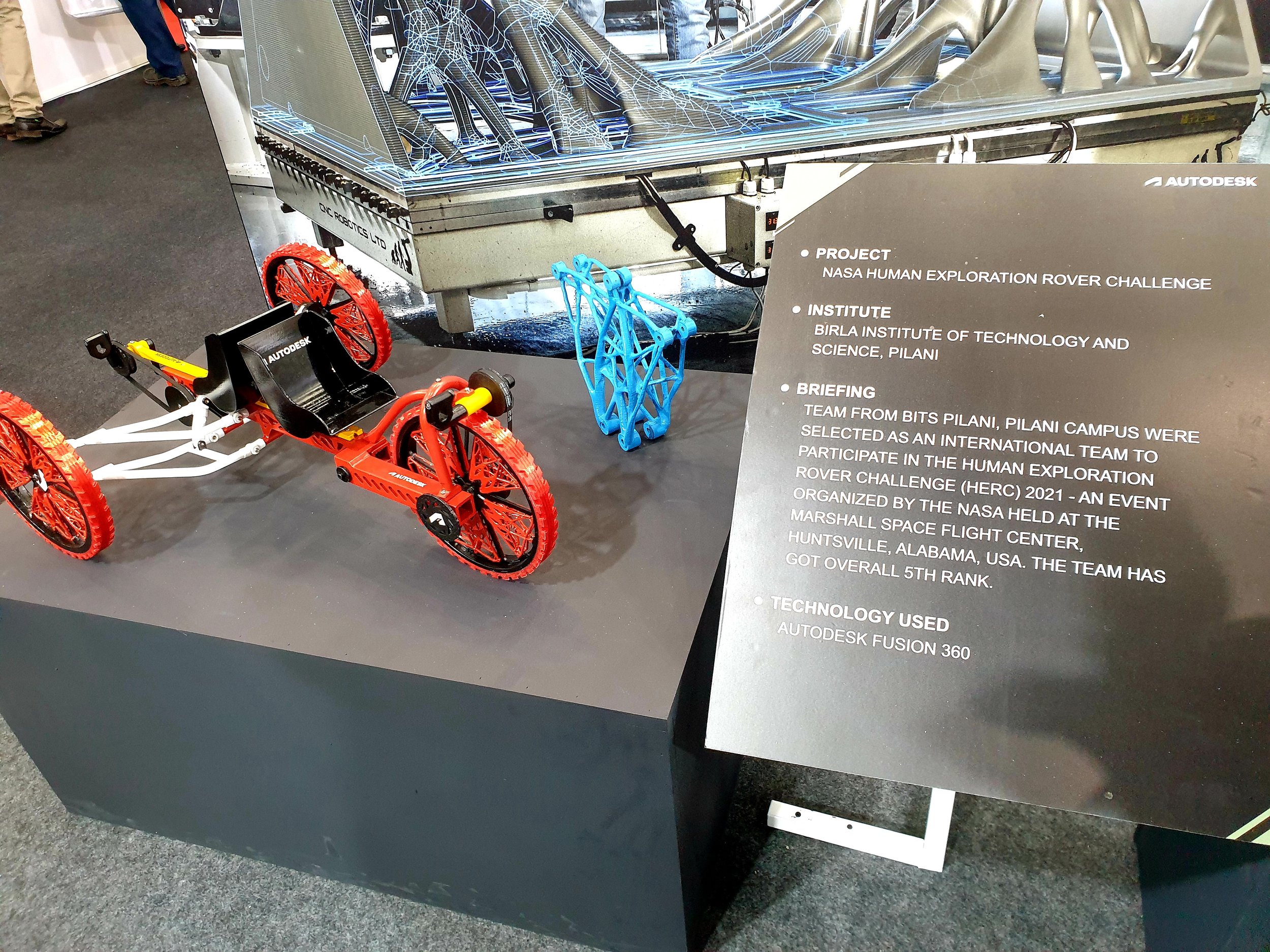 A model of a rover vehicle with red and black parts on display next to a sign detailing the NASA Human Exploration Rover Challenge participation by the Birla Institute of Technology and Science, Pilani. The sign mentions the use of Autodesk Fusion 36