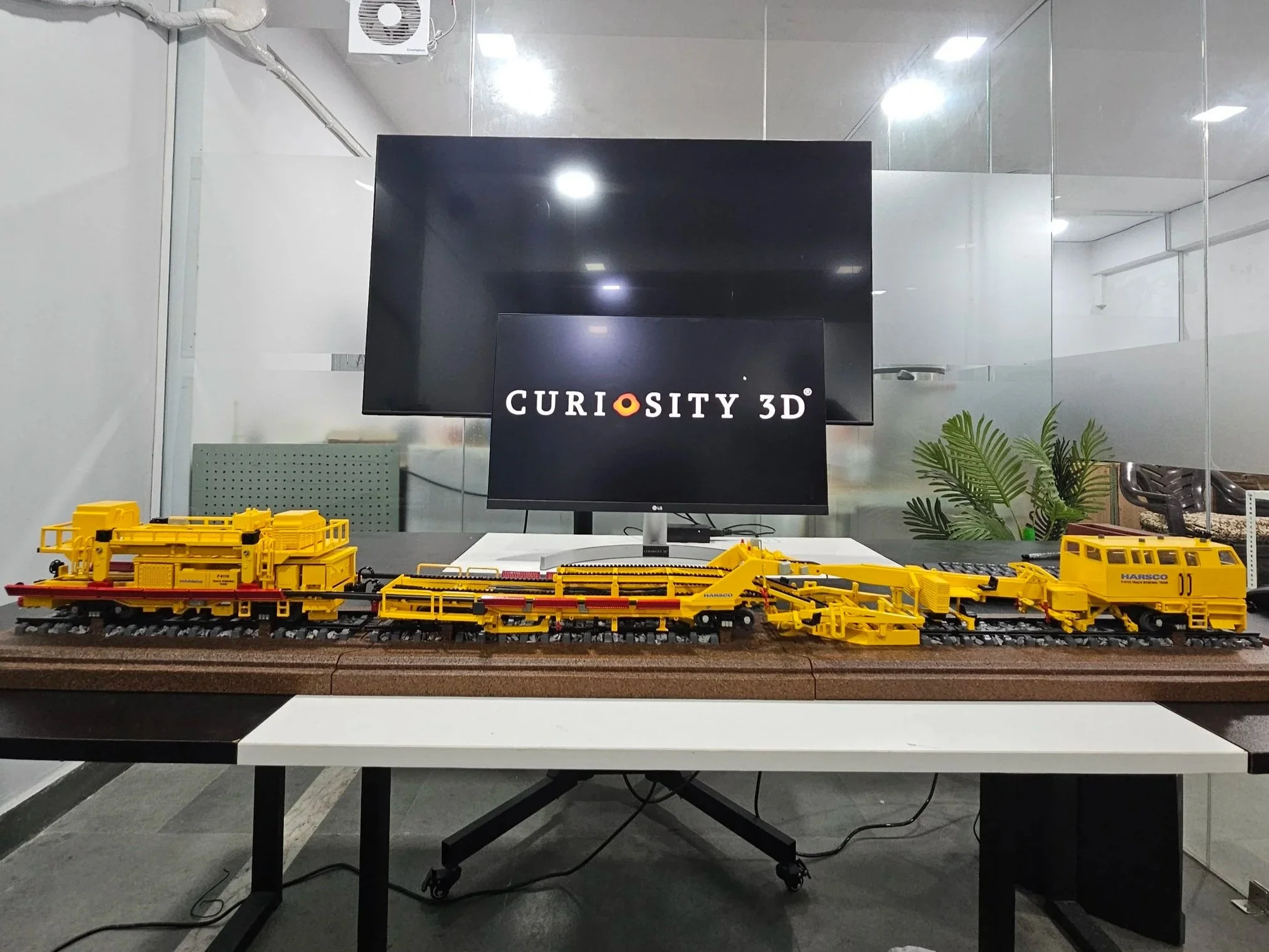Table with yellow LEGO train models and a computer monitor displaying a logo that reads "CURIOSITY 3D" in an office or lab setting with glass partitions and a potted plant.