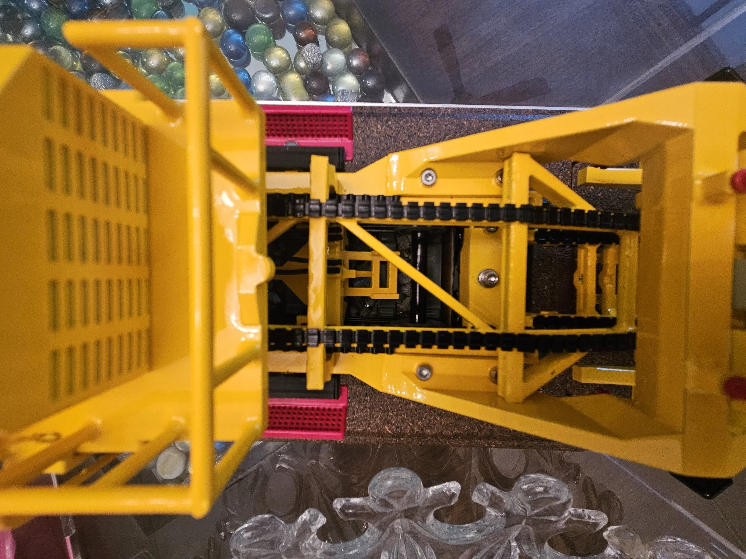 A yellow shopping cart with black wheels, positioned above a pile of clear circular plastic caps or lids, with a background of multicolored marbles on a dark surface.