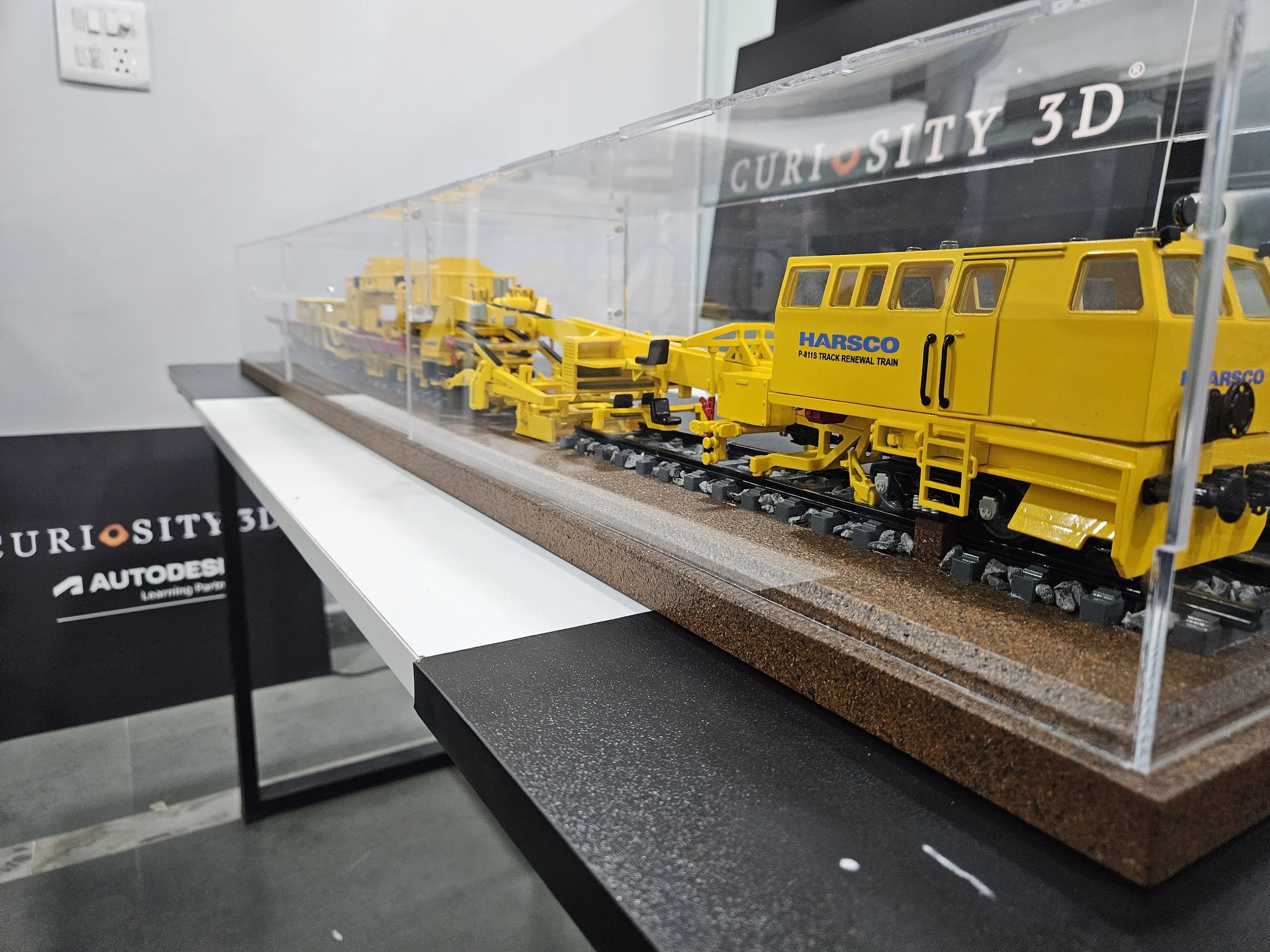 A detailed model of a yellow track renewal train inside a protective clear display case at a museum or exhibit.