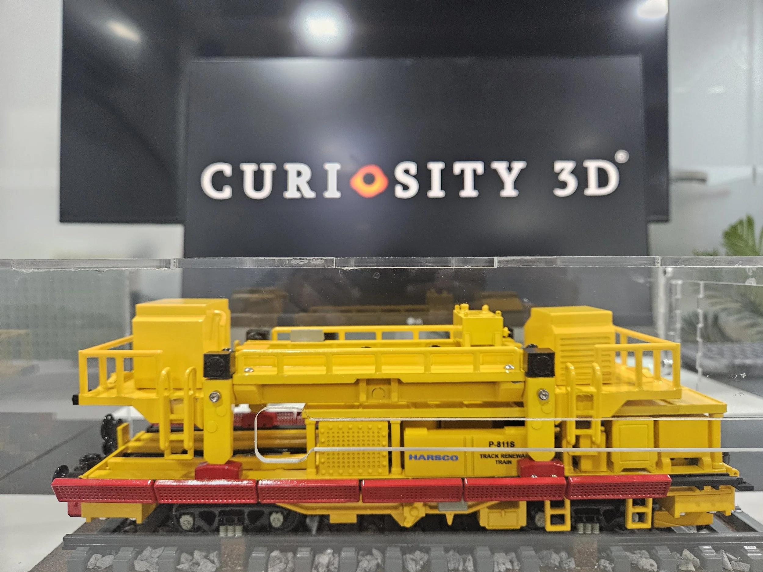 Model train tracks with yellow and red train cars in front of a black display sign that reads 'CURIOSITY 3D' with a circular orange logo in the middle.