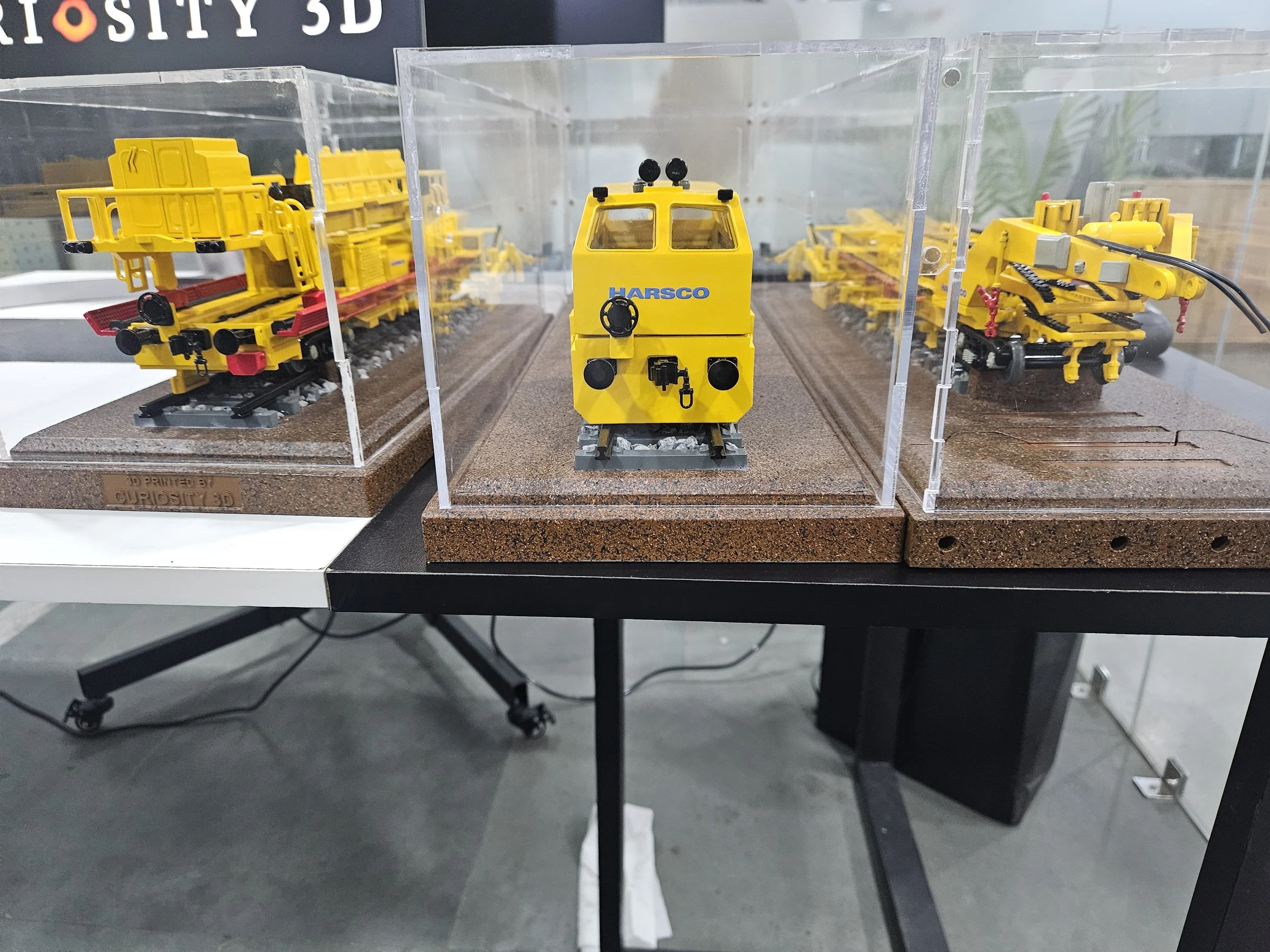 A display of yellow model construction vehicles, including a crane and a rail vehicle, placed on a textured base with a transparent divider in a showroom or exhibit setting.