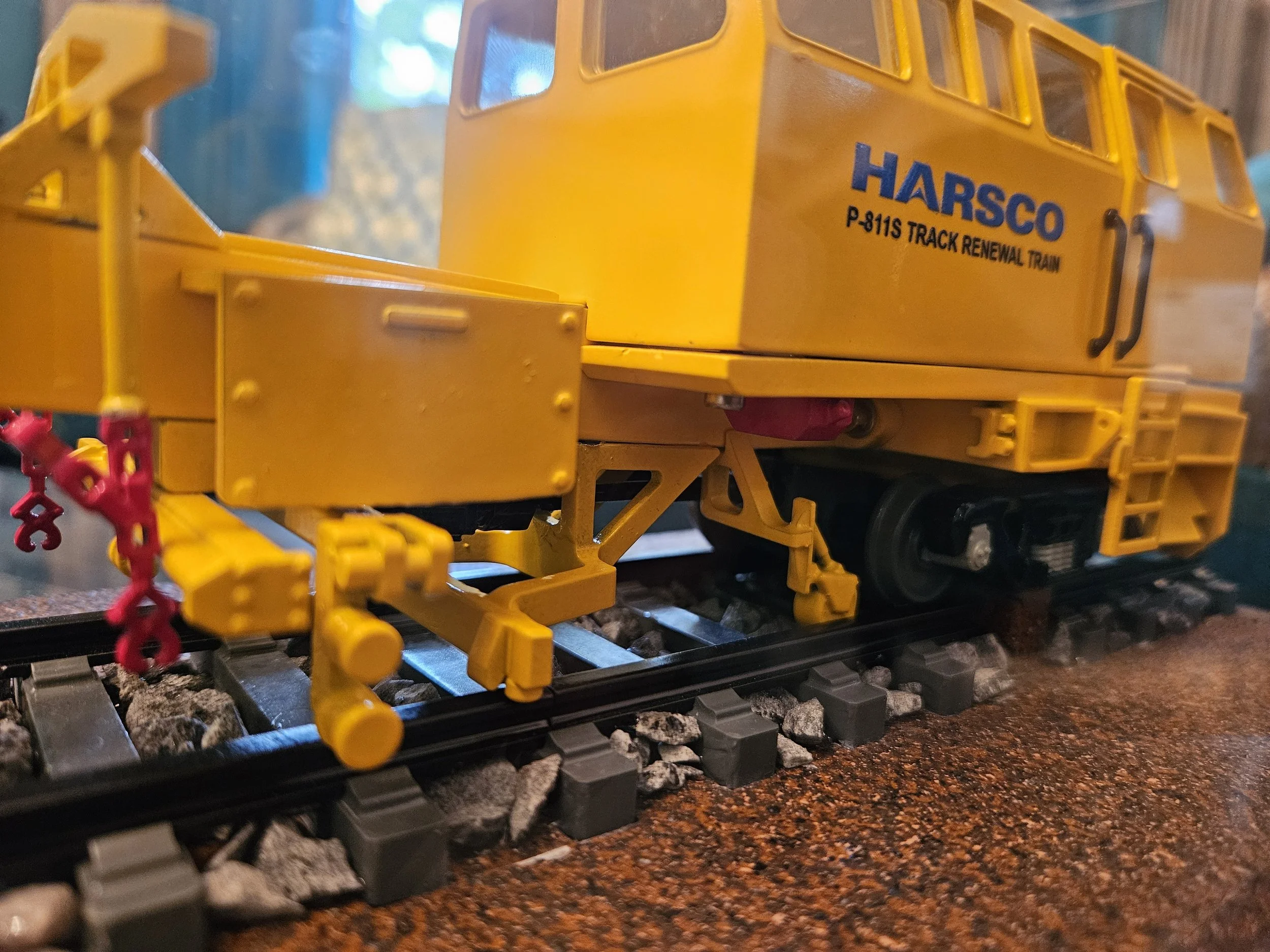 A yellow toy train car with the text 'HARSCO P-811S TRACK RENEWAL TRAIN' printed on its side, on model train tracks with gravel and a blurred background.