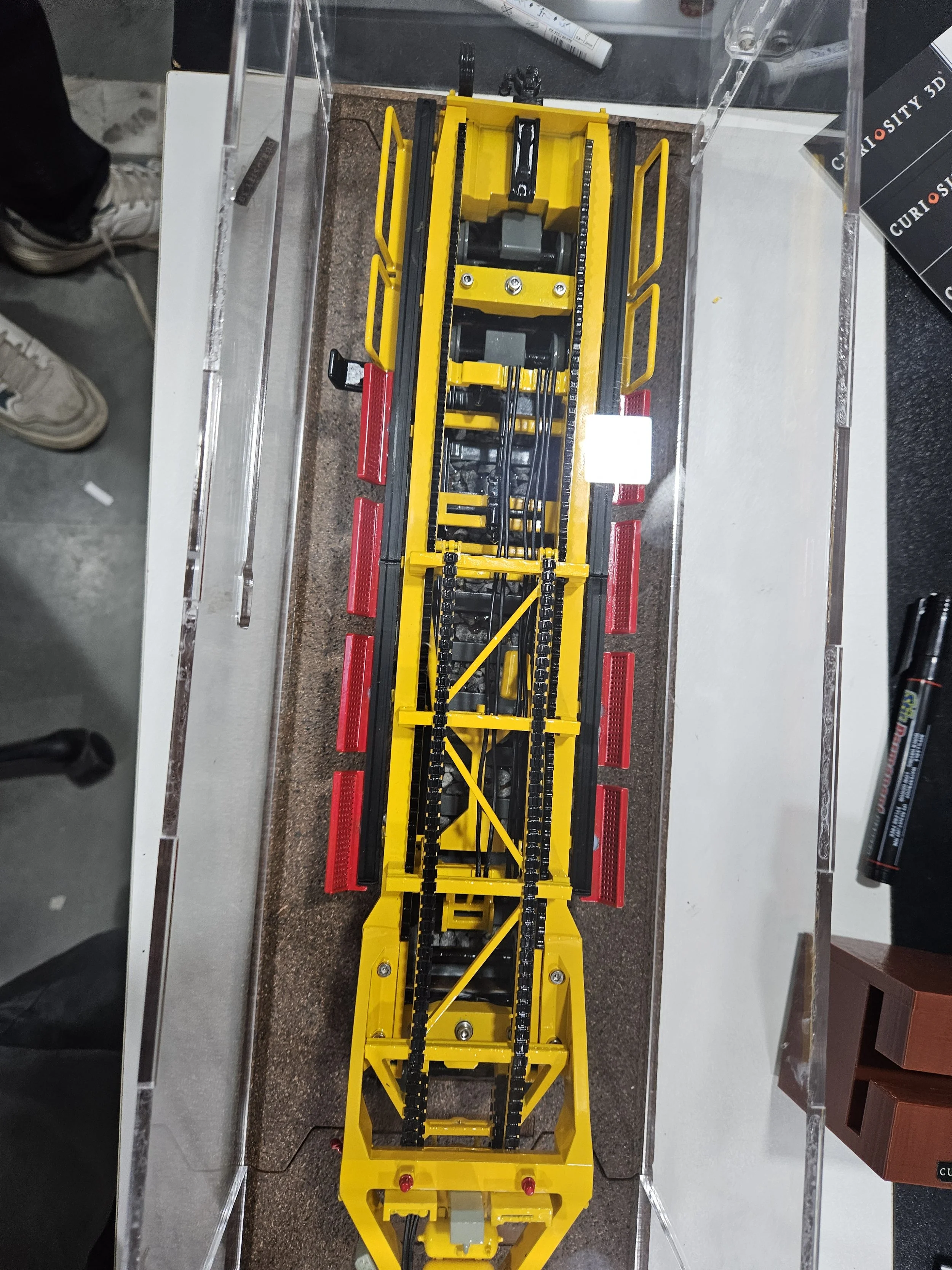 View of a yellow mechanical model or machine with black and gray parts, displayed on a table with various objects and papers around it.