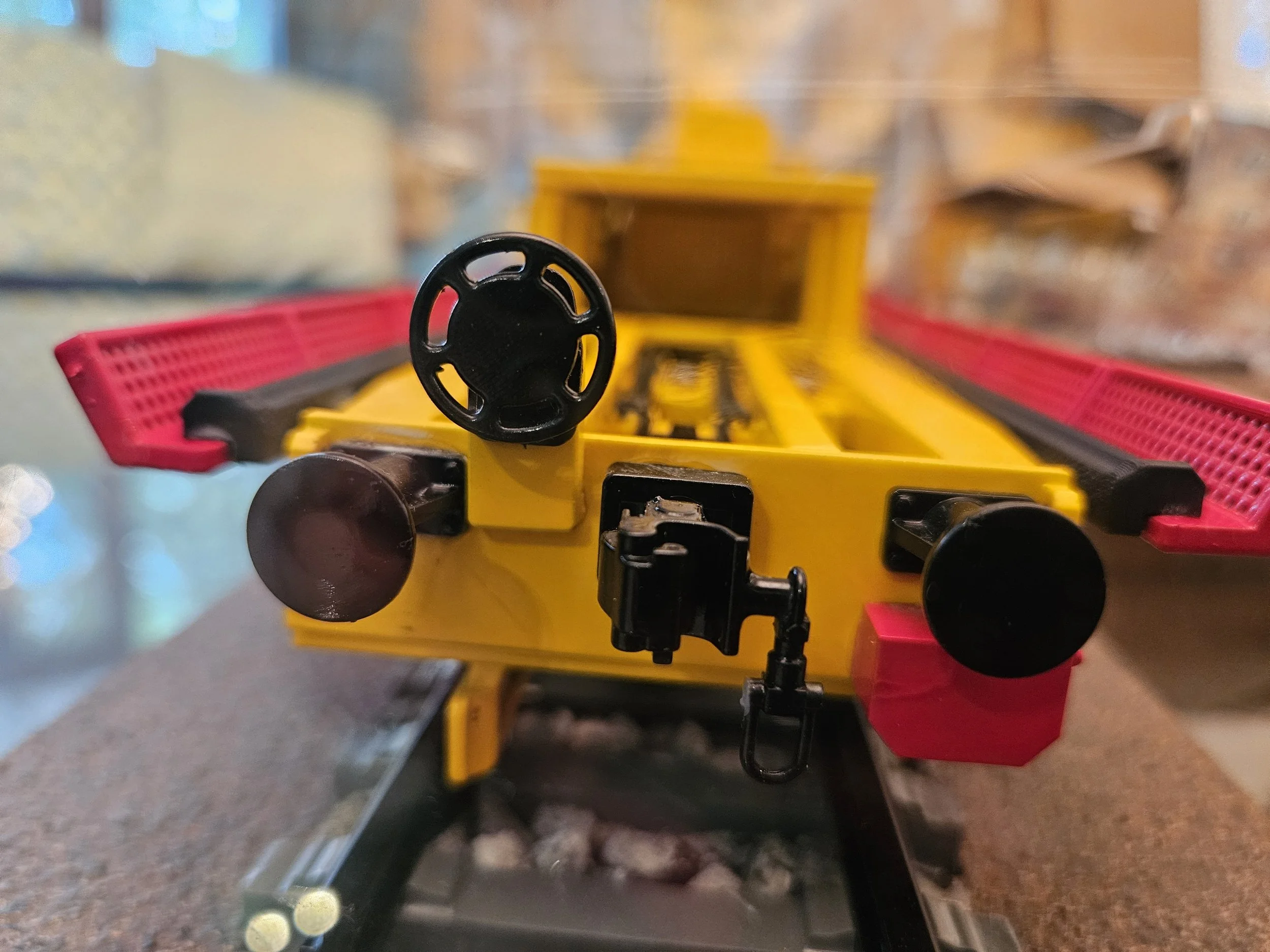 Close-up of a yellow toy bulldozer with a black steering wheel and red safety rails, showing the machine from the front with a blurred background.