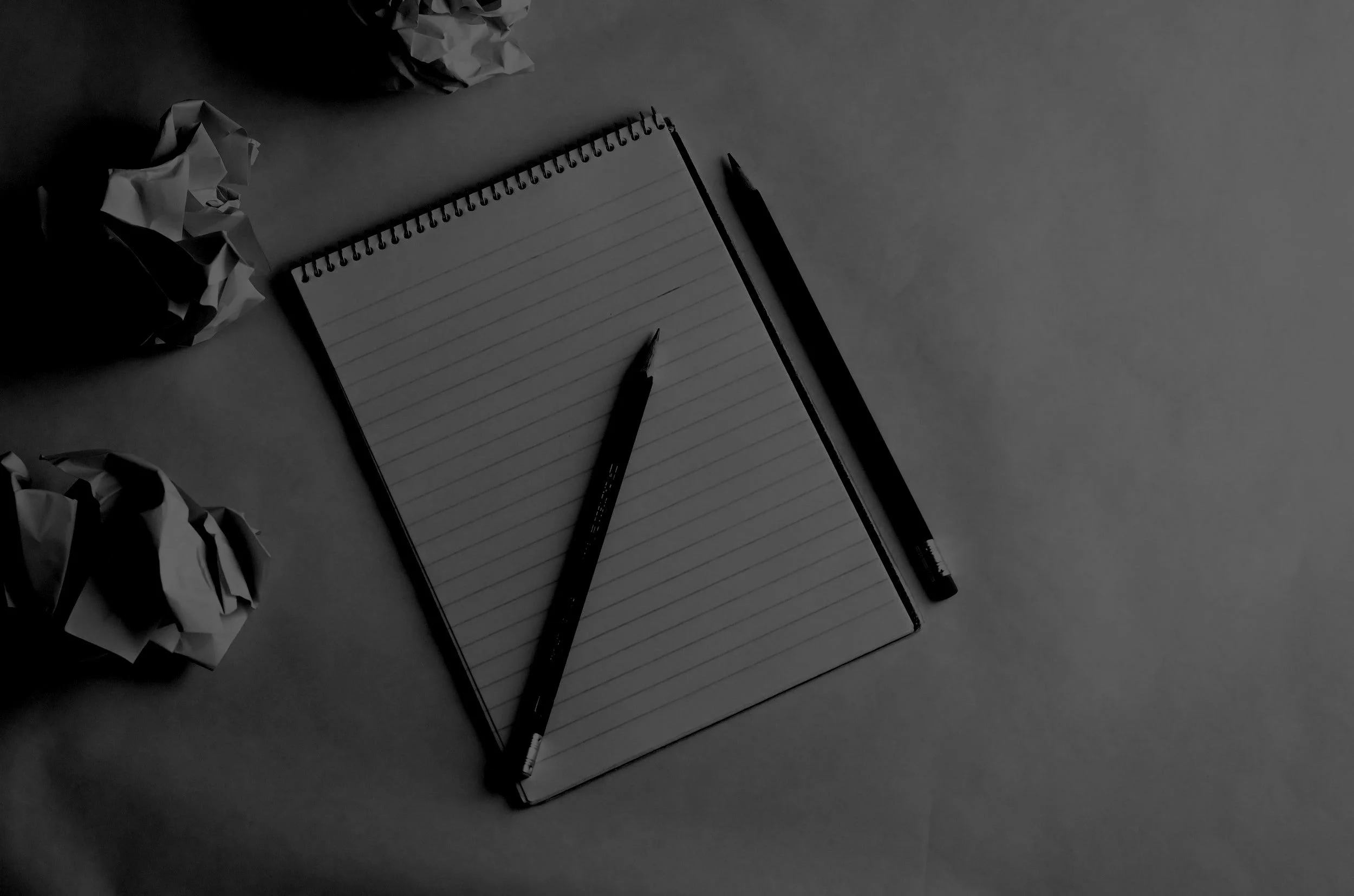 Black and white image of a lined spiral notebook with a pencil on top, another pencil beside it, and crumpled papers around.