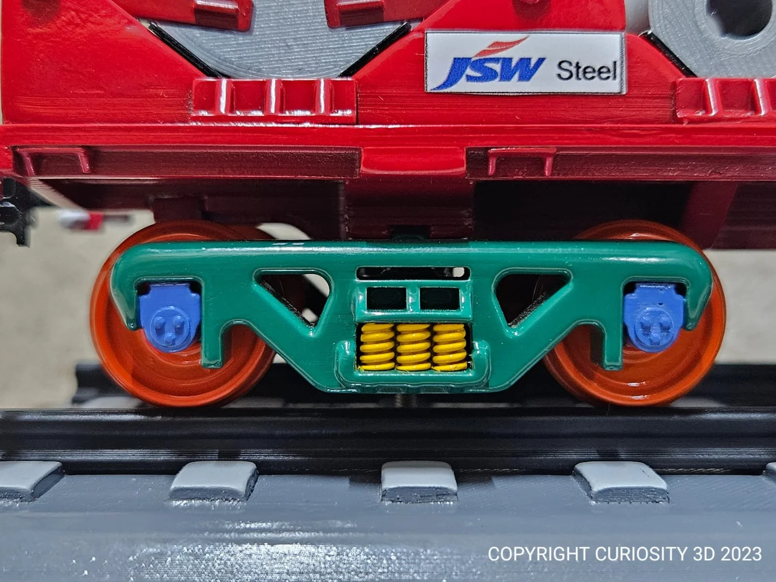 Close-up of a toy train set featuring colorful wheels and a visible "JSW Steel" logo. The train has bright orange wheels, a green frame, and blue and yellow details, positioned on a toy track.