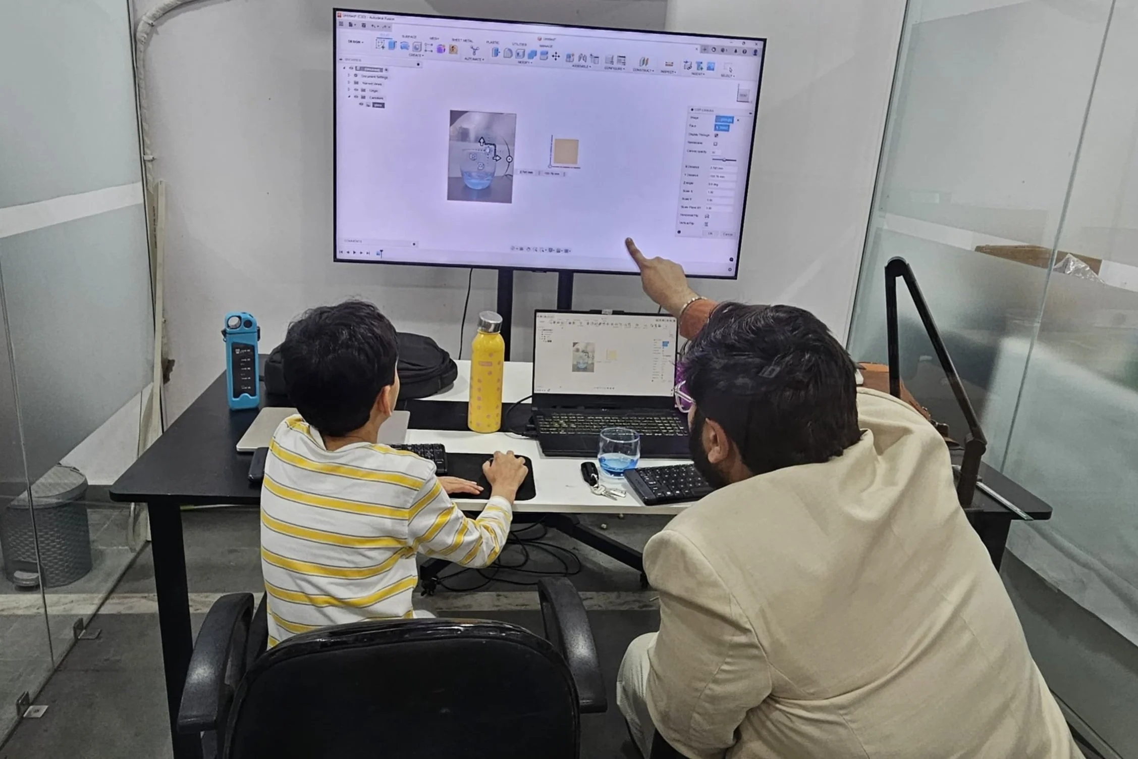 A man and a child working on a computer with a large monitor displaying design software, situated in an office setting with a yellow water bottle on the desk.