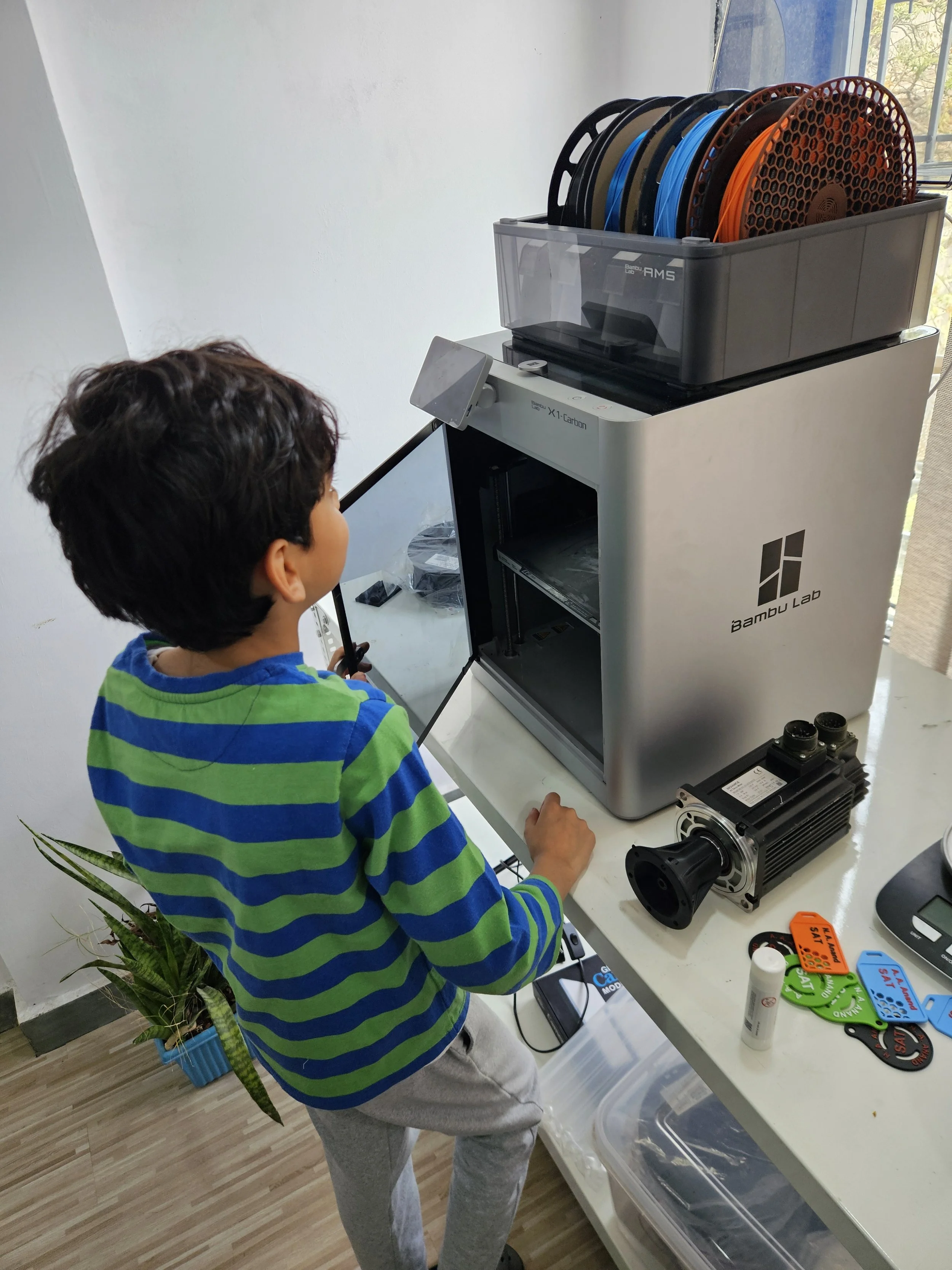 A child wearing a green and blue striped shirt standing in front of a Bambu Lab X1 Carbon 3D printer with multiple filament spools on top. The setup includes various tools on the table and a plant nearby.