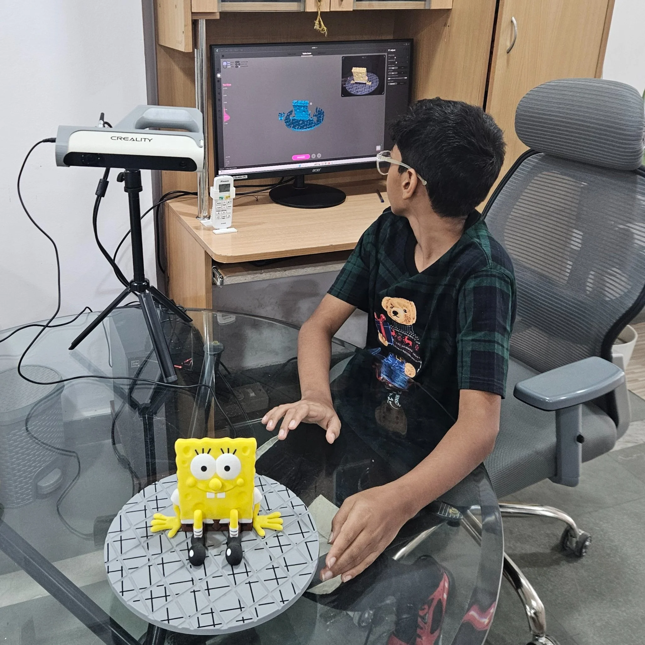 A child sitting at a desk working with a 3D printer and computer, alongside a 3D model of a cartoon character on a turntable.