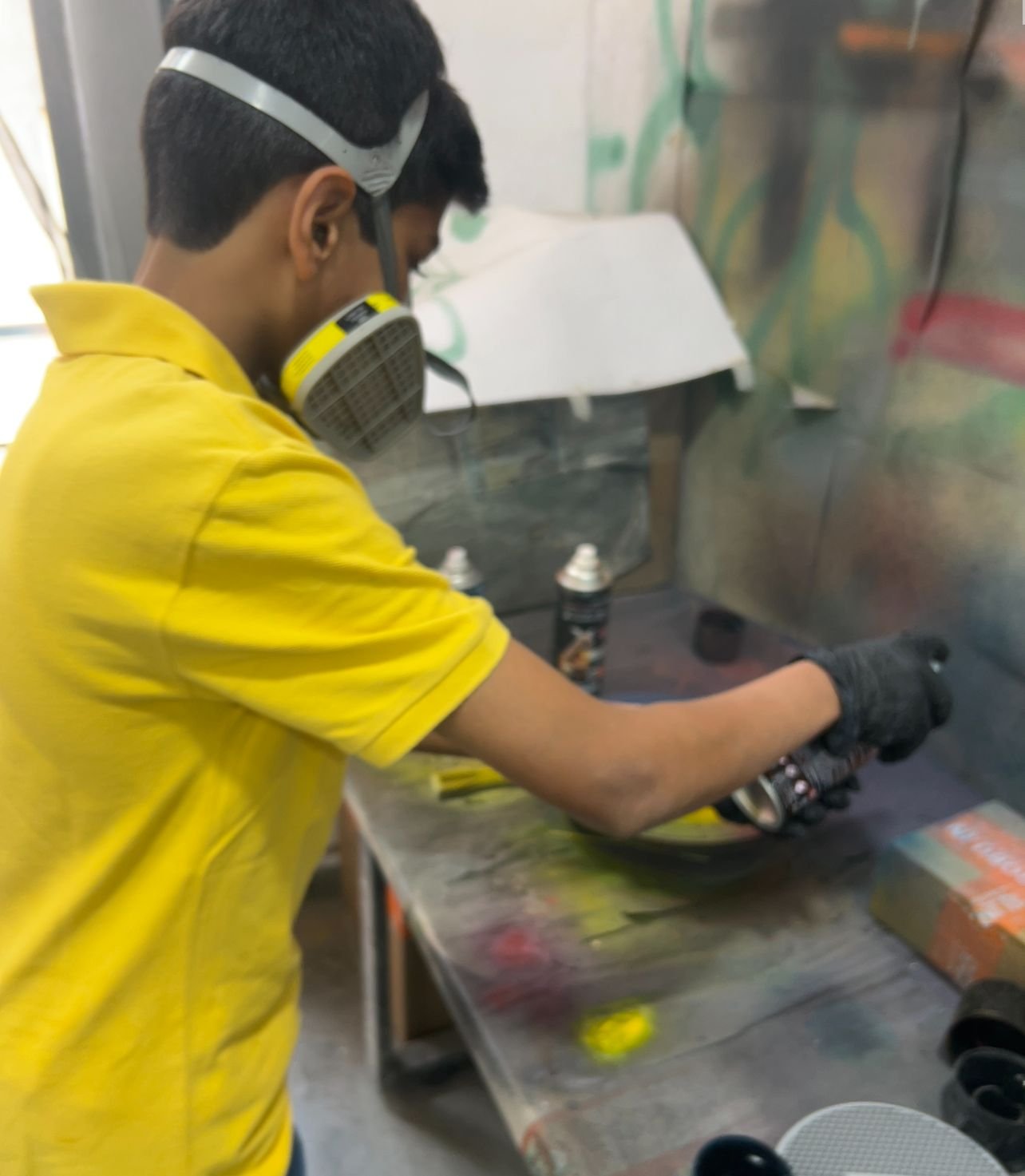 Person wearing a yellow shirt, black gloves, and a respirator mask, spray painting on a table in an artist's studio with spray paint cans and graffiti artwork in the background.
