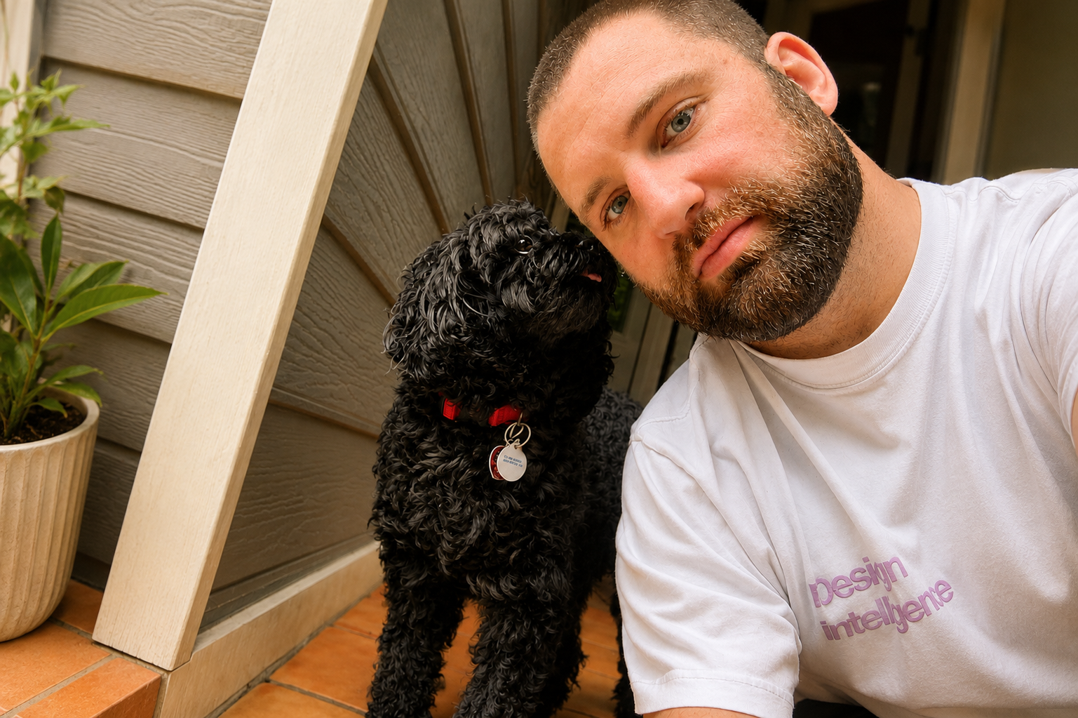 A man with a beard taking a selfie with a curly black dog on a porch.