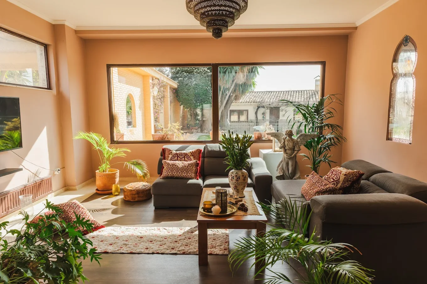 Sala de estar con sillones grises, plantas, una mesa de madera con decoración y una ventana grande que deja entrar luz natural.