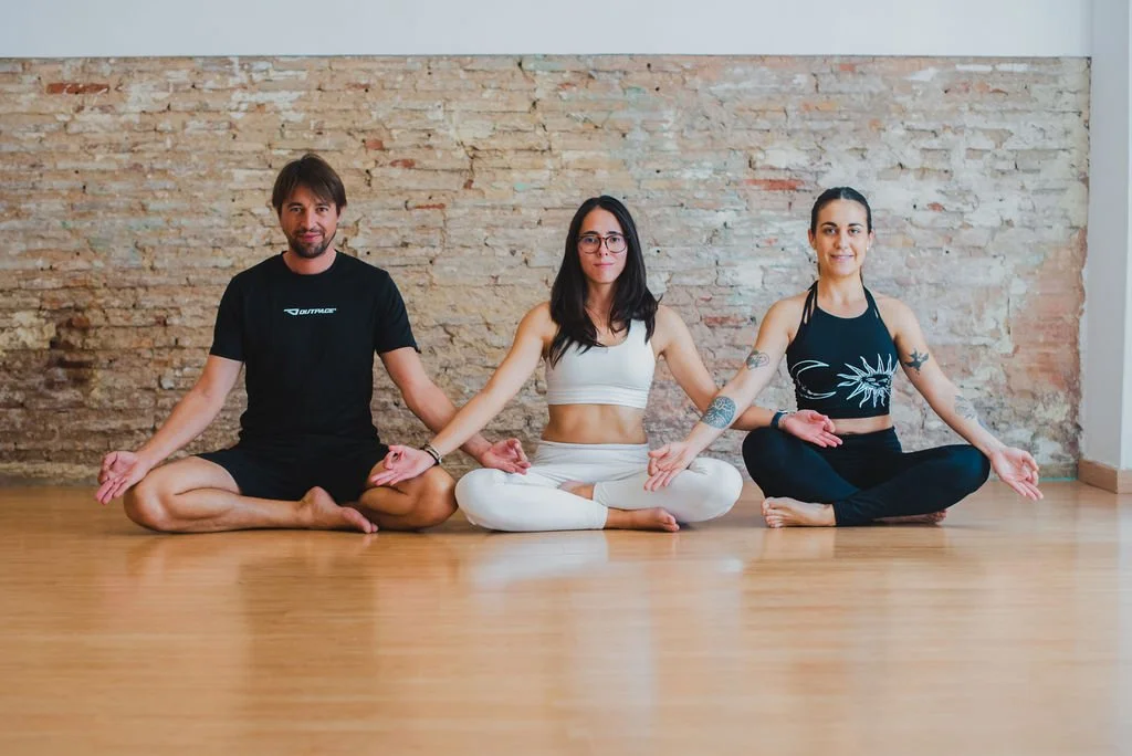 Tres personas practicando yoga en una sala con pared de ladrillos expuestos, sentadas en posición de loto con las piernas cruzadas y las manos sobre las rodillas.