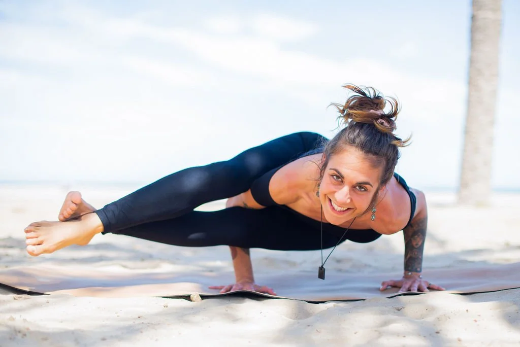 Mujer haciendo yoga en la playa, en posición de plancha lateral, sonriendo y usando ropa deportiva negra.