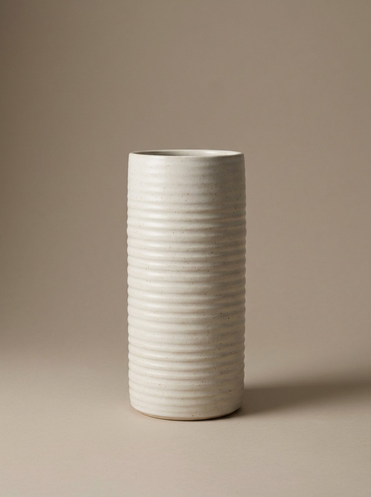 A tall, cylindrical white ceramic vase with horizontal ribbed texture, standing on a beige surface against a plain beige background.