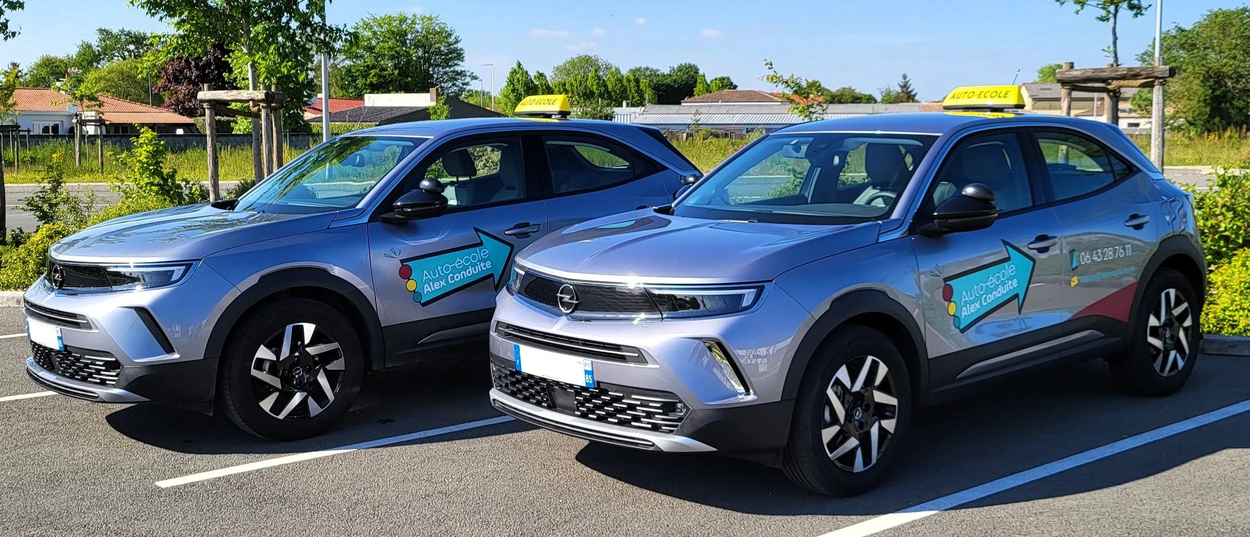 Deux voitures grises électriques stationnées dans une place de parking, avec des panneaux indiquant 'Auto-école Alex Conduite' et un taillight rouge, sous un ciel ensoleillé avec des arbres verts en arrière-plan.