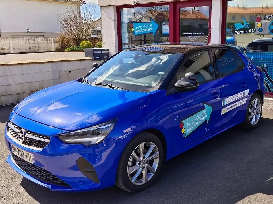 Voiture bleue avec un panneau publicitaire pour une auto-école, stationnée dehors devant un bâtiment avec large fenêtres.