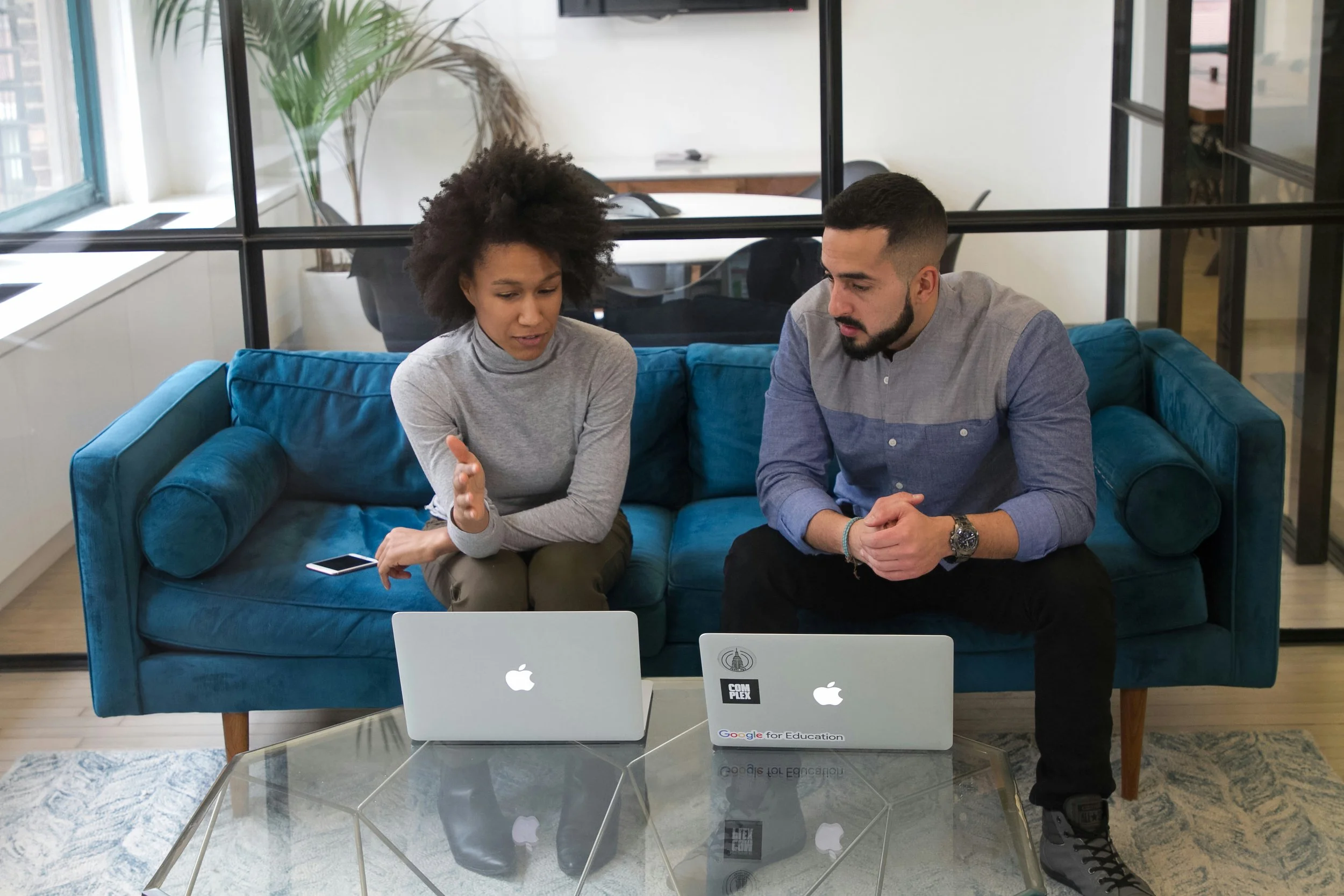 Two people sitting on a blue couch, engaged in conversation. A woman with curly dark hair wearing a gray top and a man with short dark hair and a beard wearing a gray shirt. In front of them, there are two laptops, one with an Apple logo and the other with a Google for Education and Chrome sticker. The room has large windows, a glass coffee table, and office furniture in the background.