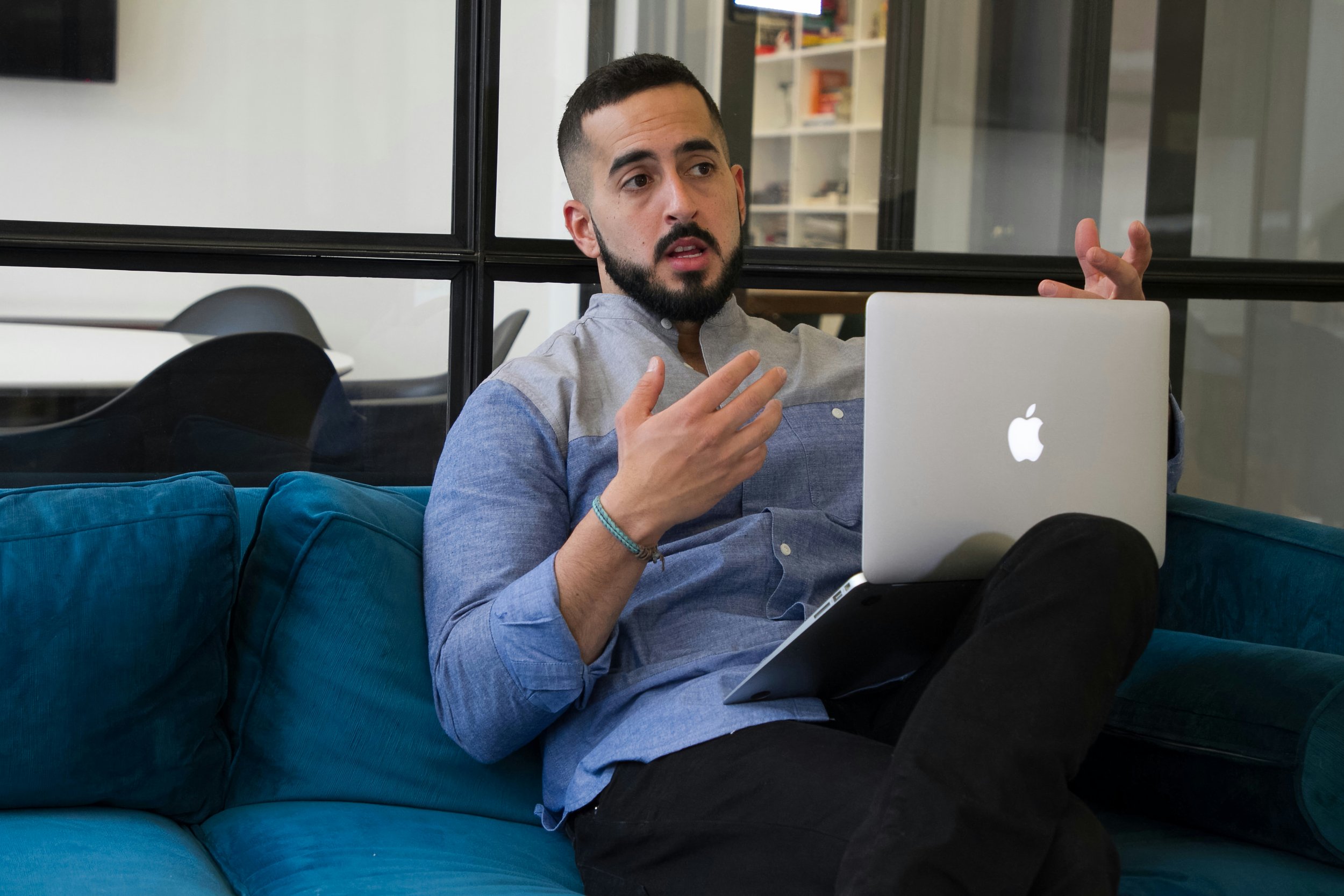 A man with a beard, short dark hair, and a two-tone shirt sitting on a blue couch using a silver MacBook laptop in a modern room with black-framed glass dividers and a bookshelf in the background.