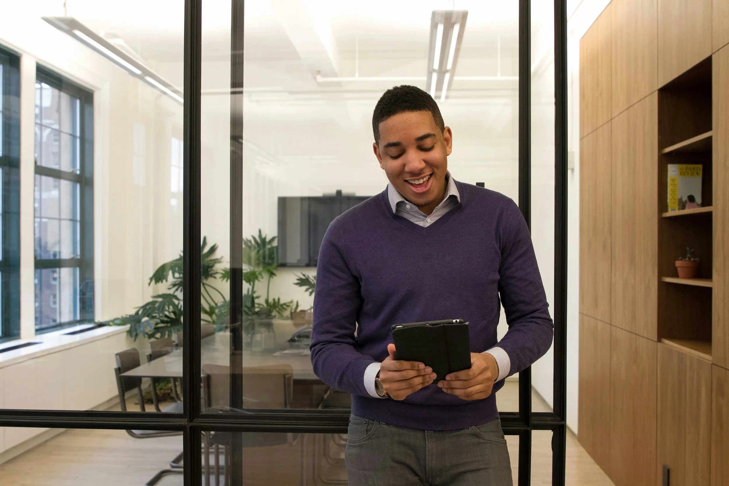 A young man in a purple sweater and gray pants standing in an office, smiling and looking at his phone.