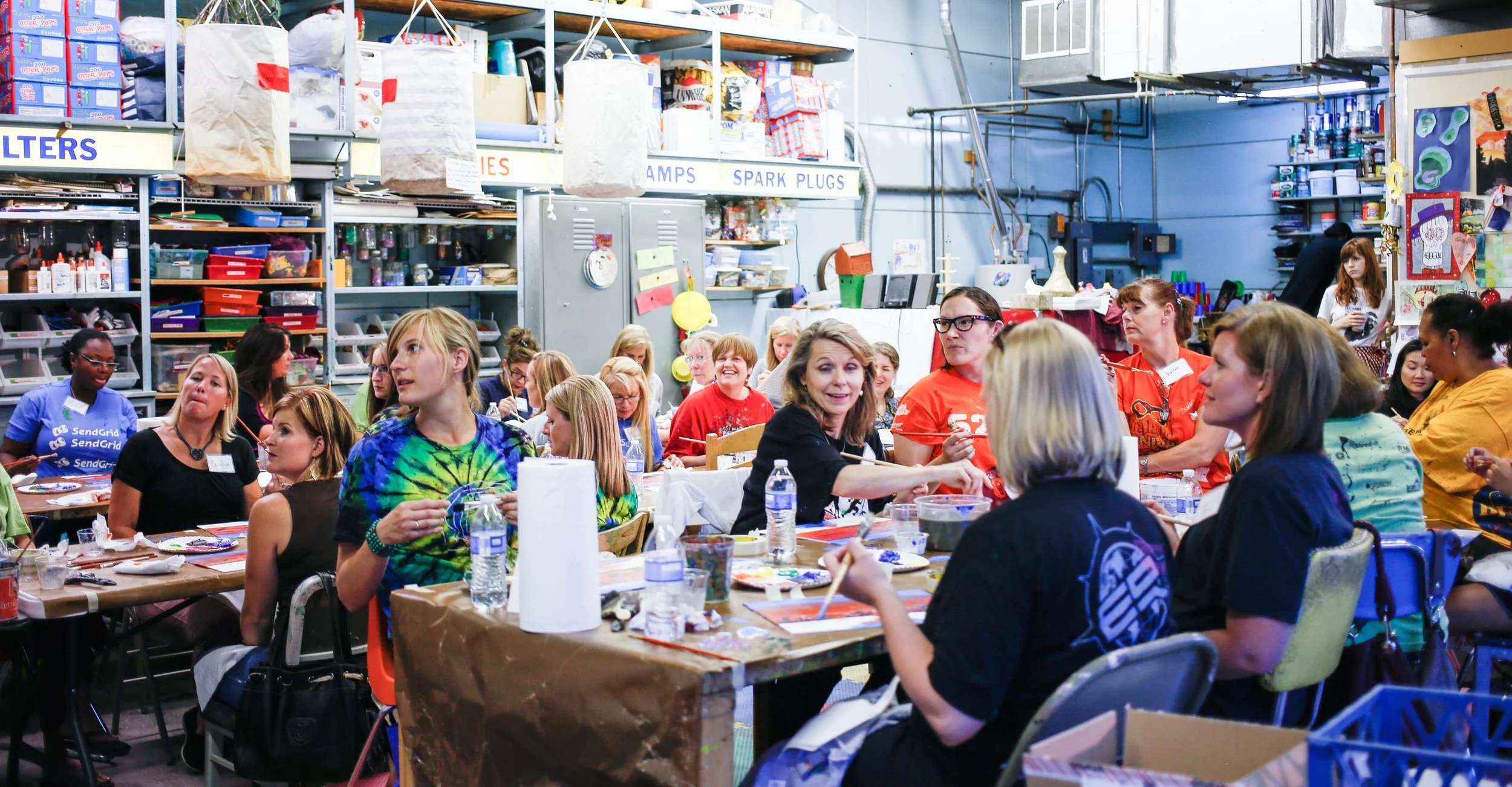 Group of people sitting around tables in a warehouse and participating in an arts and crafts event, with shelves of supplies in the background.