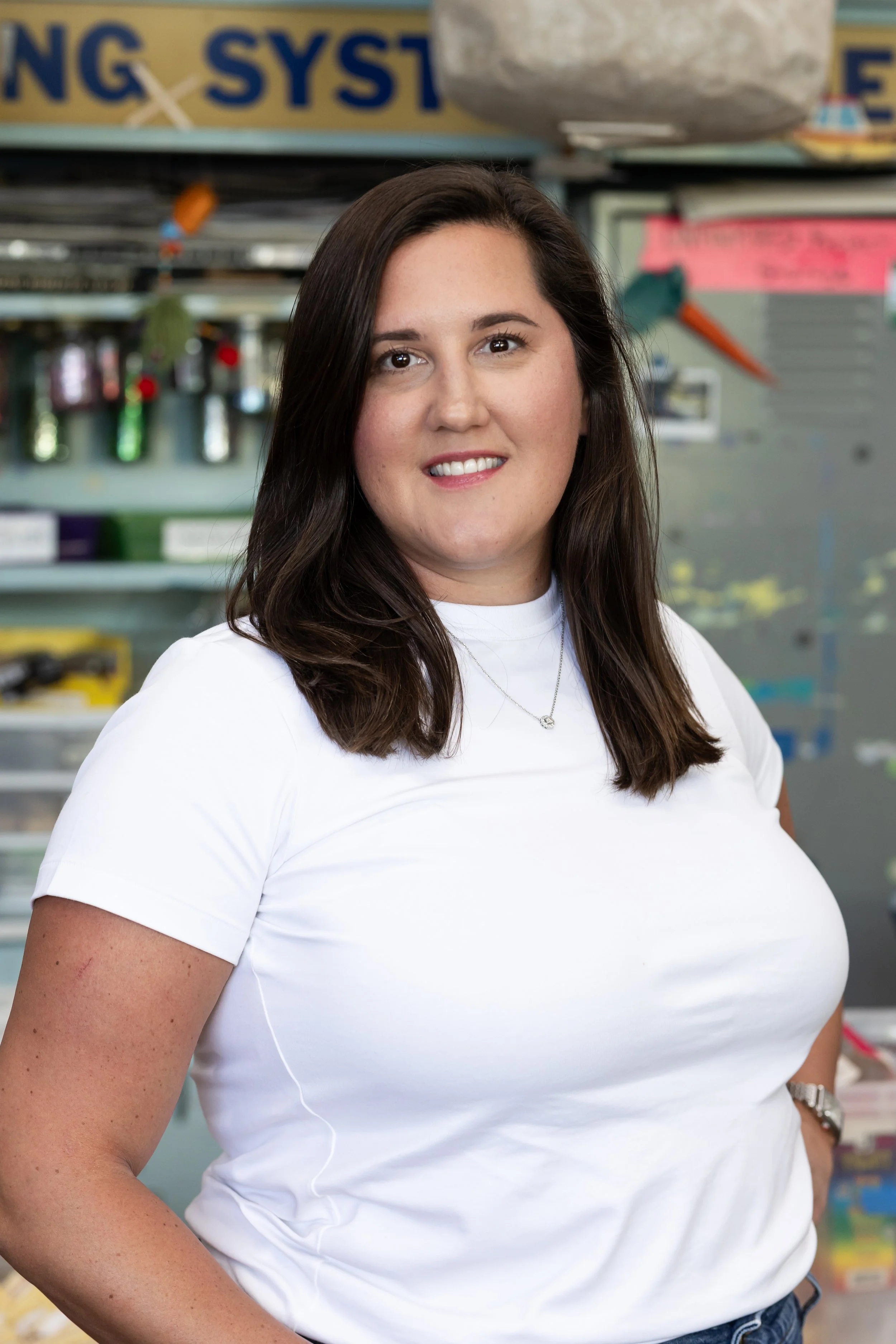 A woman with long dark hair, wearing a white t-shirt and necklace, standing in front of a store shelf with various items.