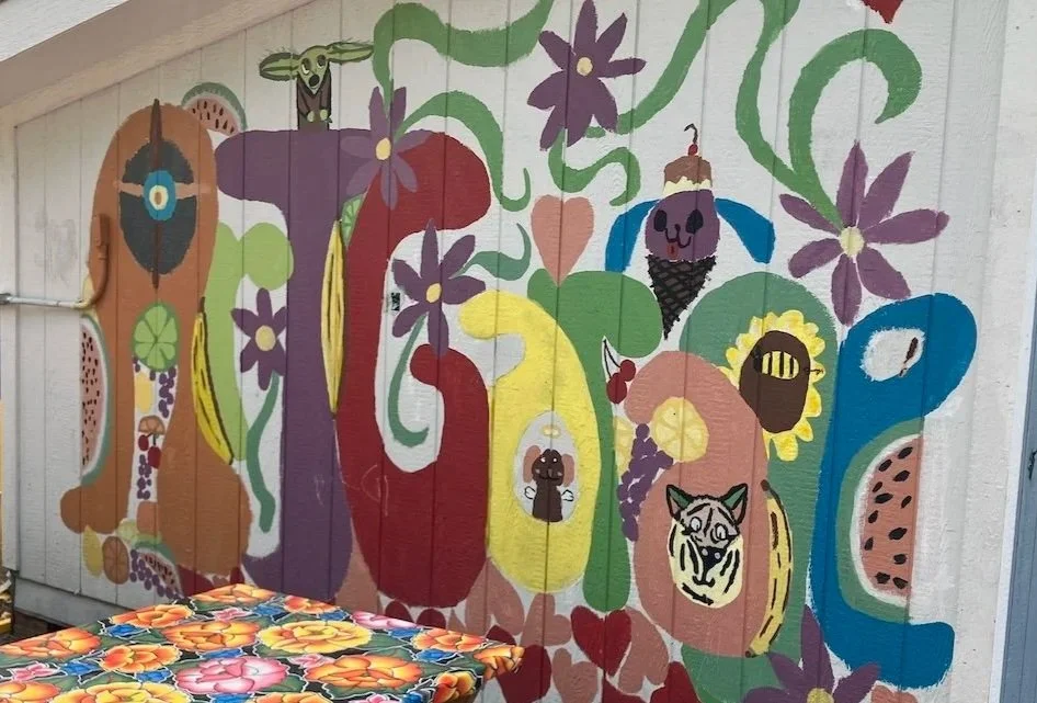 Colorful mural on a wooden wall featuring large, abstract flowers, animals like a tiger, butterfly, bee, and a bird, with hanging strings and paper cutouts.