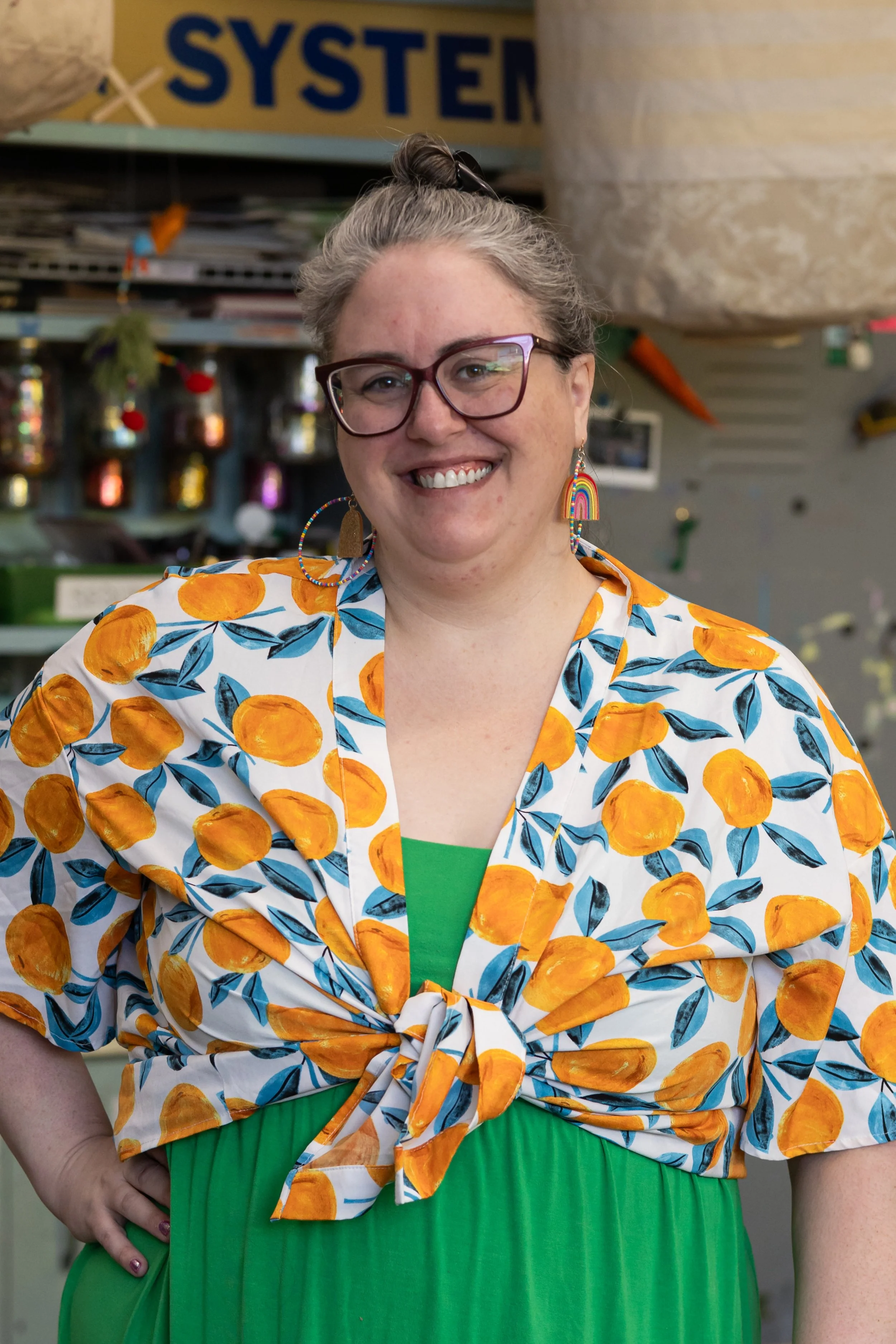 A woman with gray hair in a bun wearing glasses, colorful rainbow earrings, a white shirt with orange fruit print, and a green skirt. She is smiling with her hand on her hip, standing in front of a shelf with plants and decorative items.