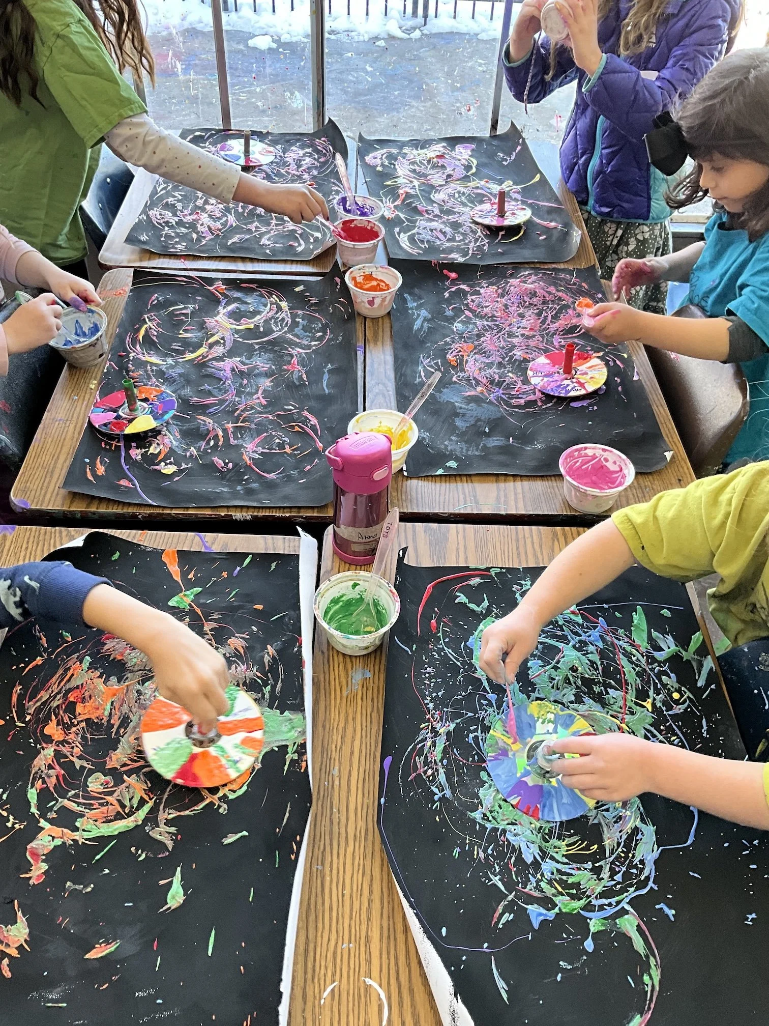 Children creating colorful abstract art with paint and spinning paper plates on black paper mats at a table.