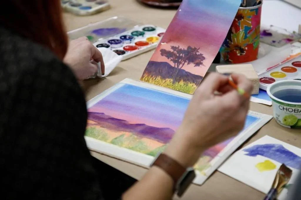 Person painting a scenic landscape with mountains, sky, and a tree using watercolors at a table with art supplies.
