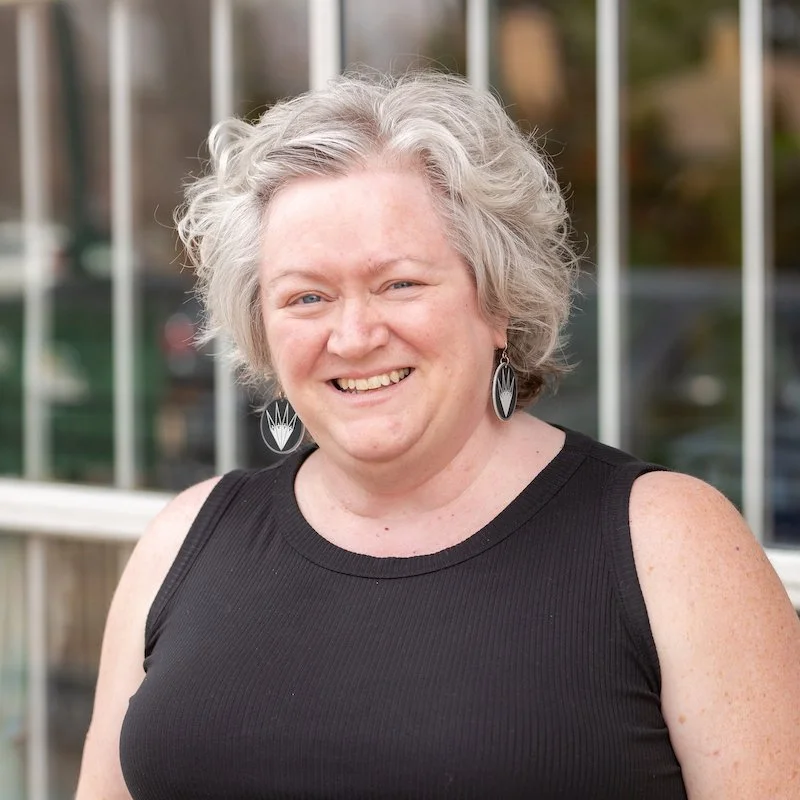A woman with curly gray hair smiling outdoors, wearing a black sleeveless top and distinctive earrings.
