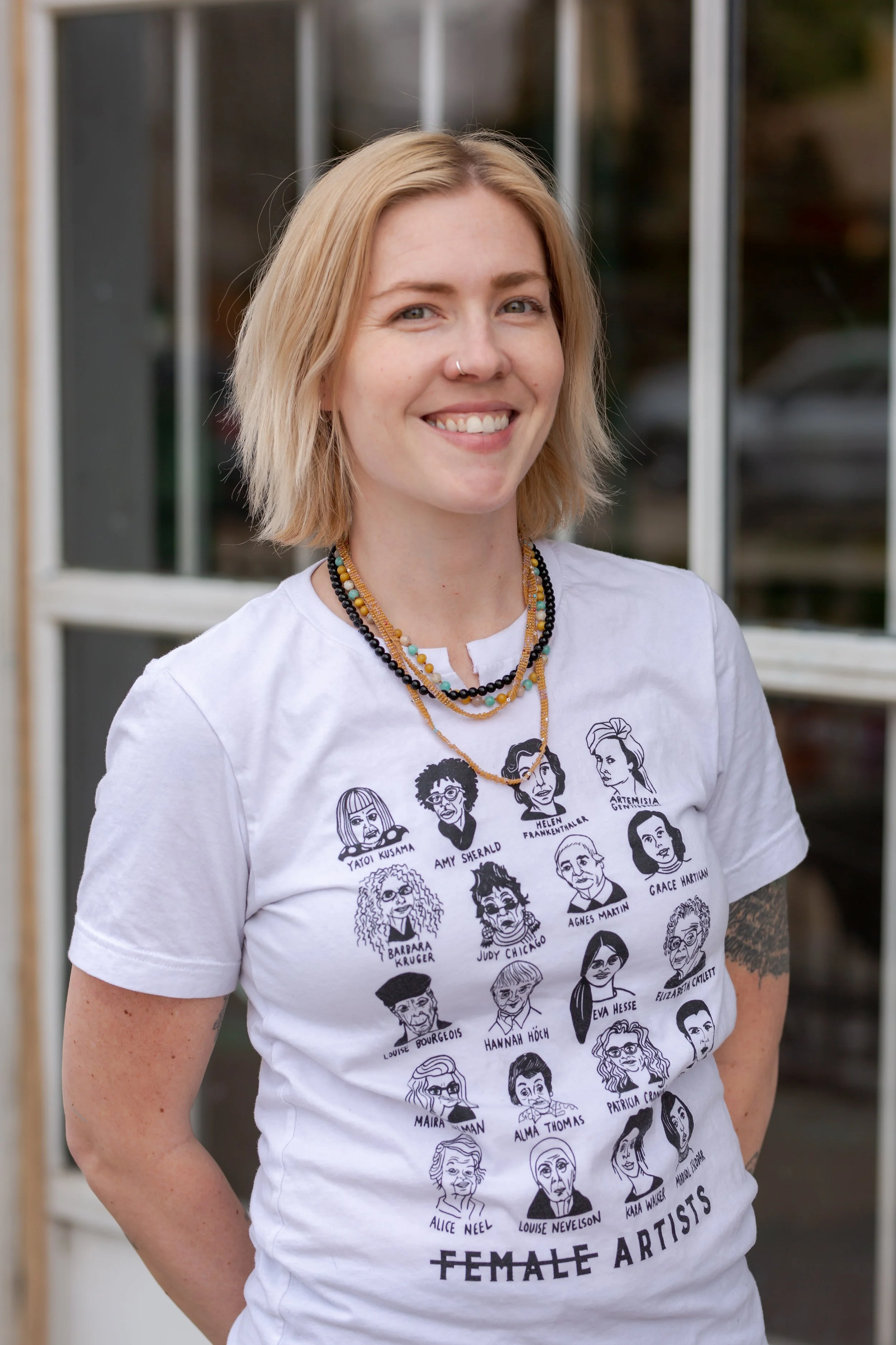 A woman with blonde hair, wearing a white T-shirt with illustrations of various female artists and their names, standing outdoors near a building with glass windows.