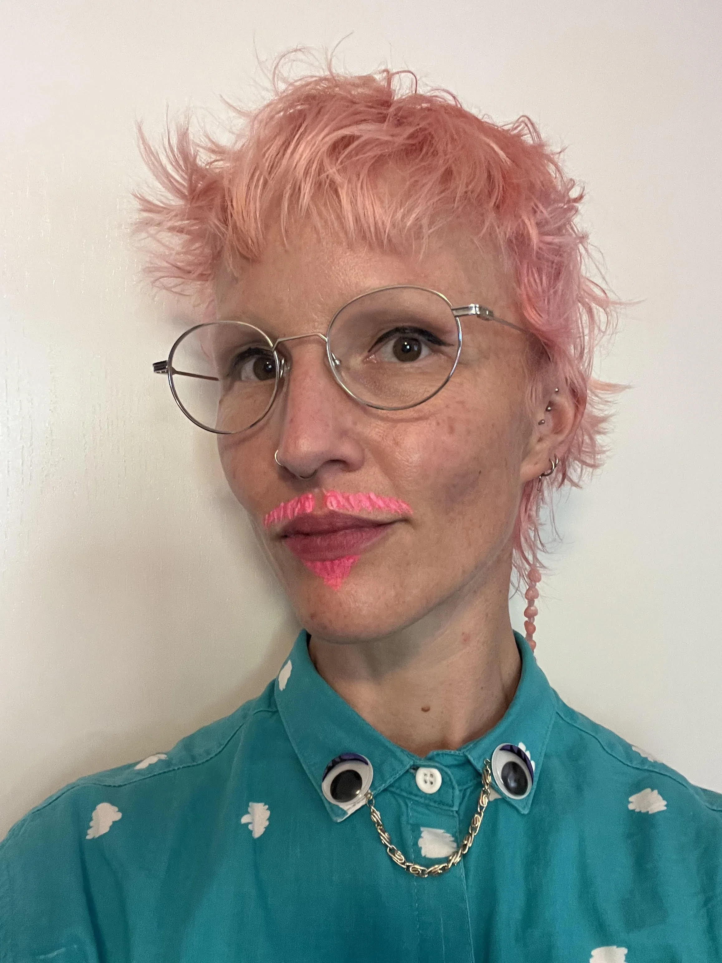 Person with short curly pink hair wearing round glasses, a blue shirt with cloud patterns, and a necklace with eye-shaped pendants.