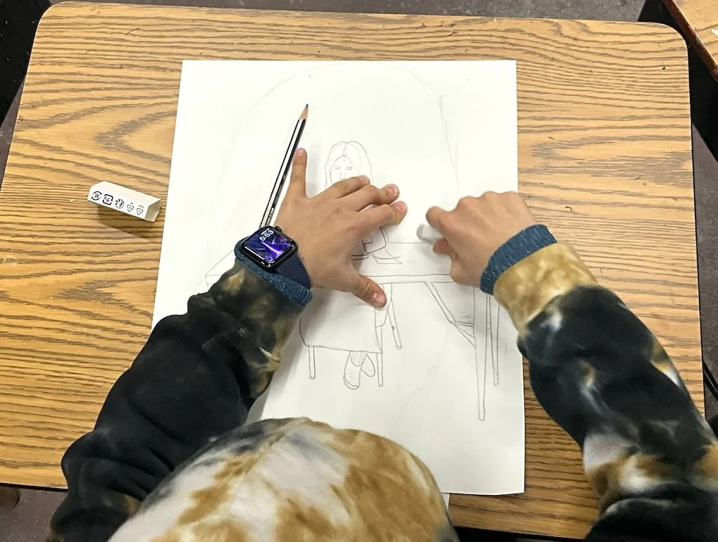 A person with a watch on their left wrist drawing a picture of a girl sitting at a table on a piece of paper. The person is erasing or shading part of the drawing.