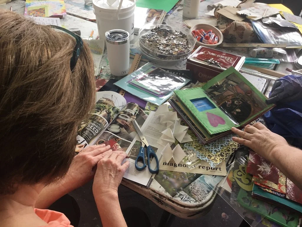 A cluttered table with crafting supplies, magazines, scissors, sequins, and various papers. Two people are working on a craft project, one cutting paper and the other opening a decorated photo album.