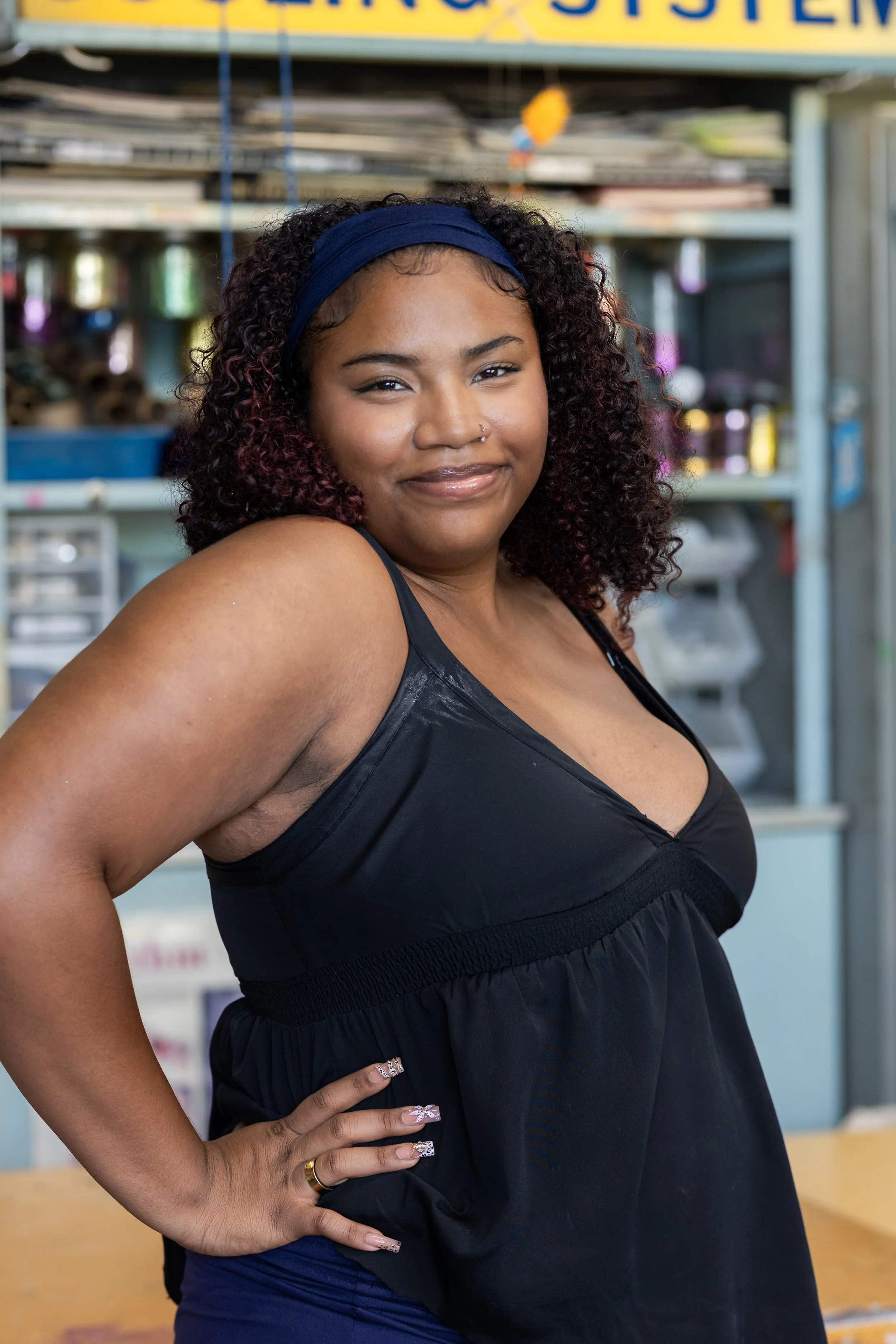 A woman with curly hair wearing a blue headband and a black sleeveless top, standing indoors with shelves of various items in the background.